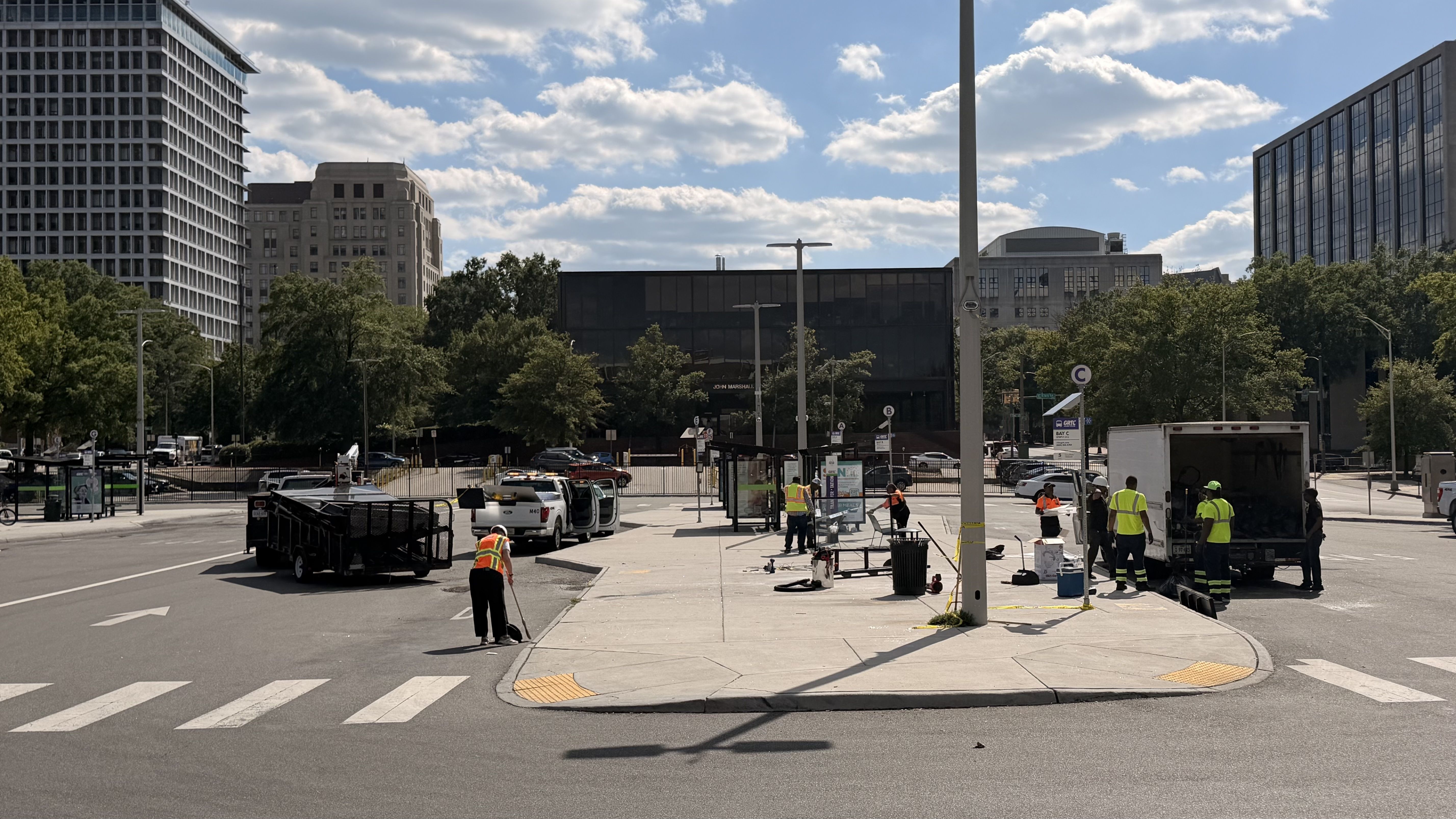 Urban street scene with construction workers in high-visibility vests near a black glass building, tall office towers, crosswalk, and a blue sky with scattered clouds.
