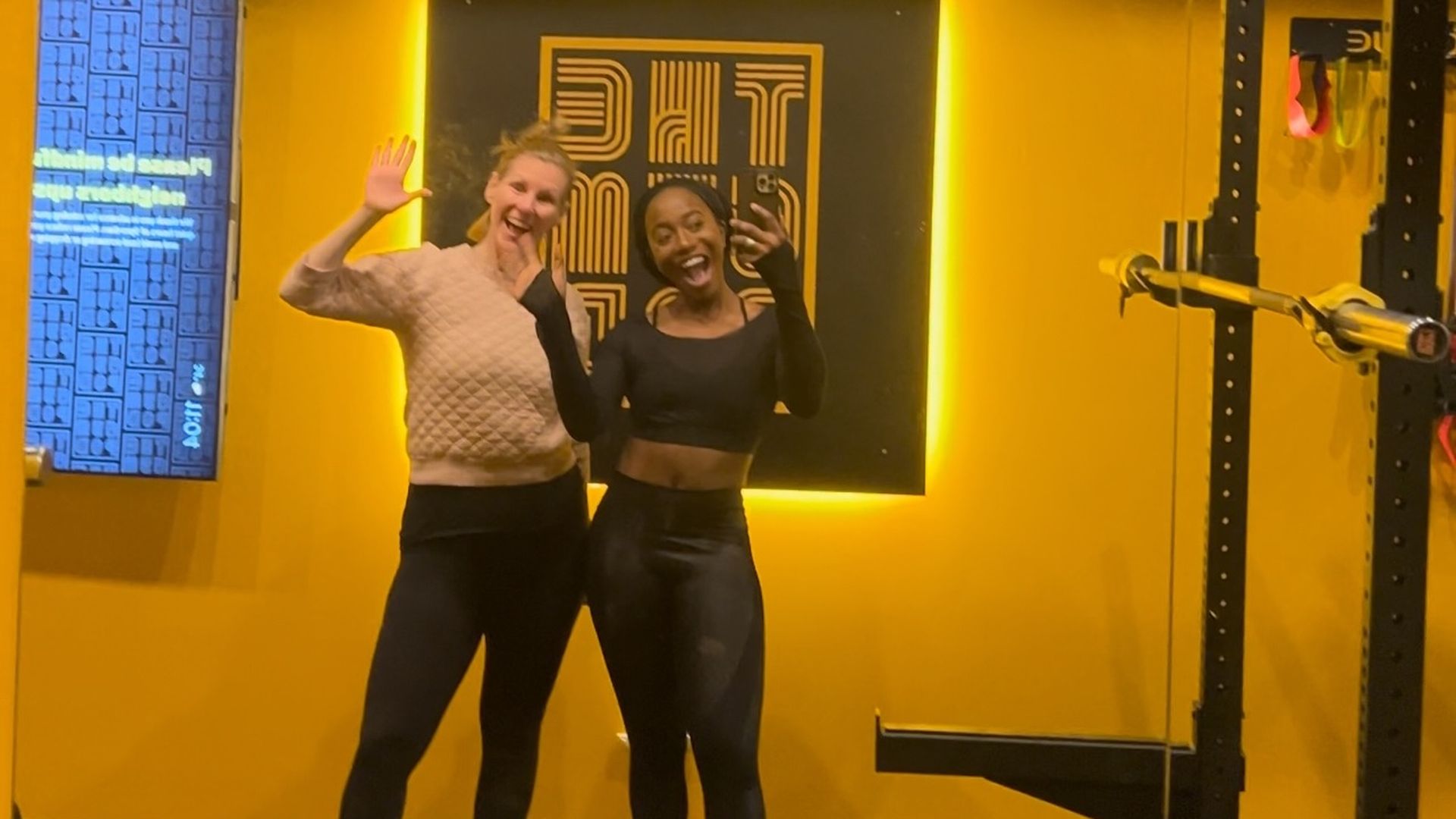 Two women wave to the camera in a gym with a yellow wall and equipment.