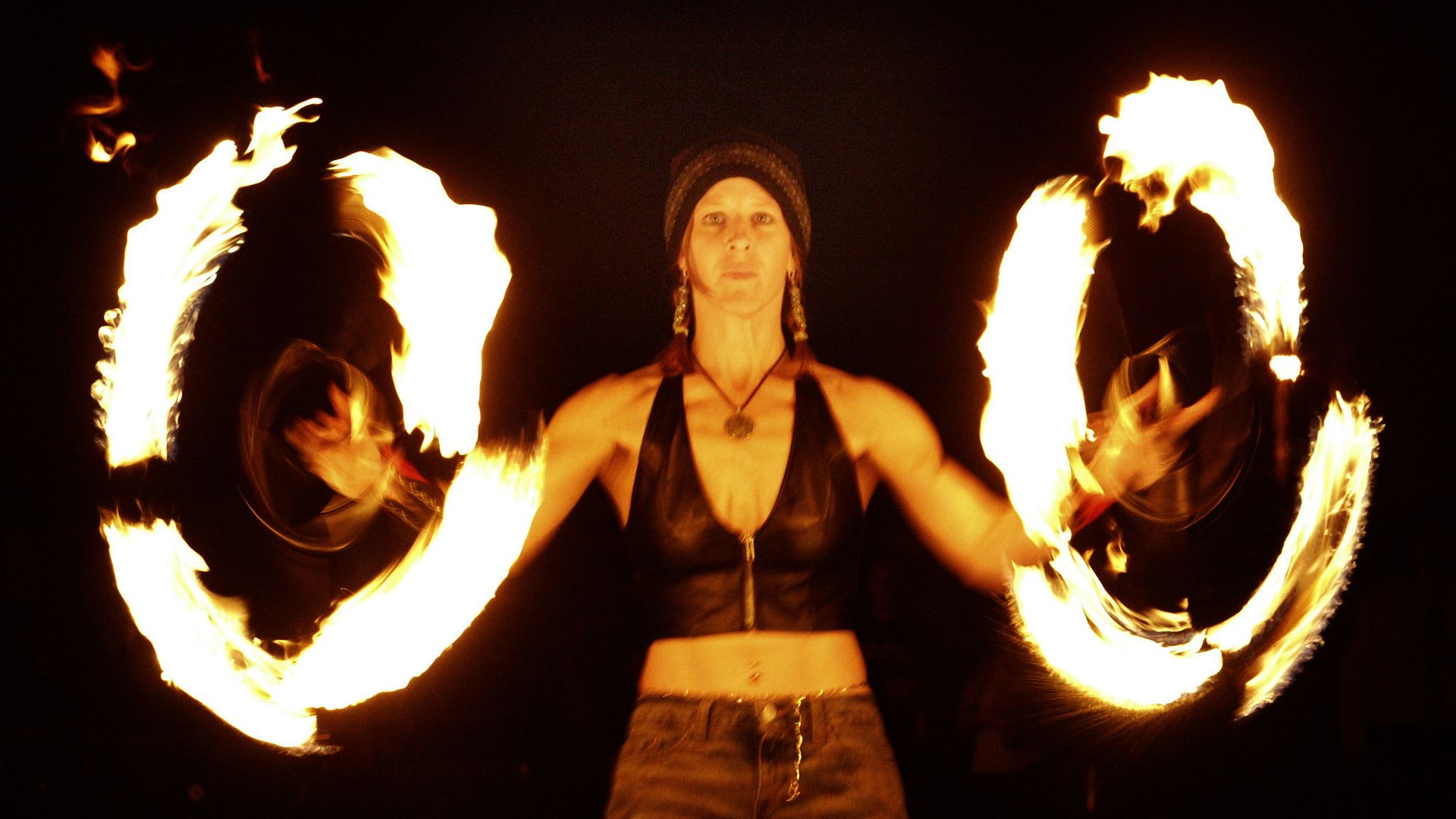 A woman wearing a knit beanie and black top spins two large rings of fire, flames arching around them in the dark.