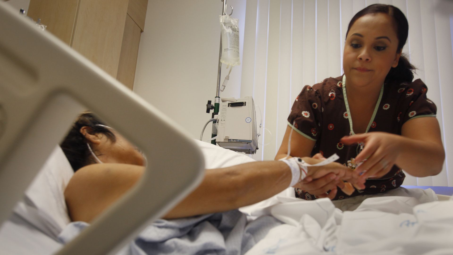 A nurse checks on an IV for a patient who is laying in a hospital bed.