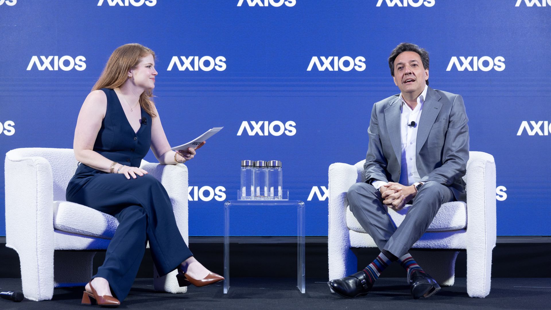 Two people on a blue Axios stage: a woman in a navy jumpsuit holds papers, and a man in a gray suit sits smiling; a small glass table with water bottles sits between them.