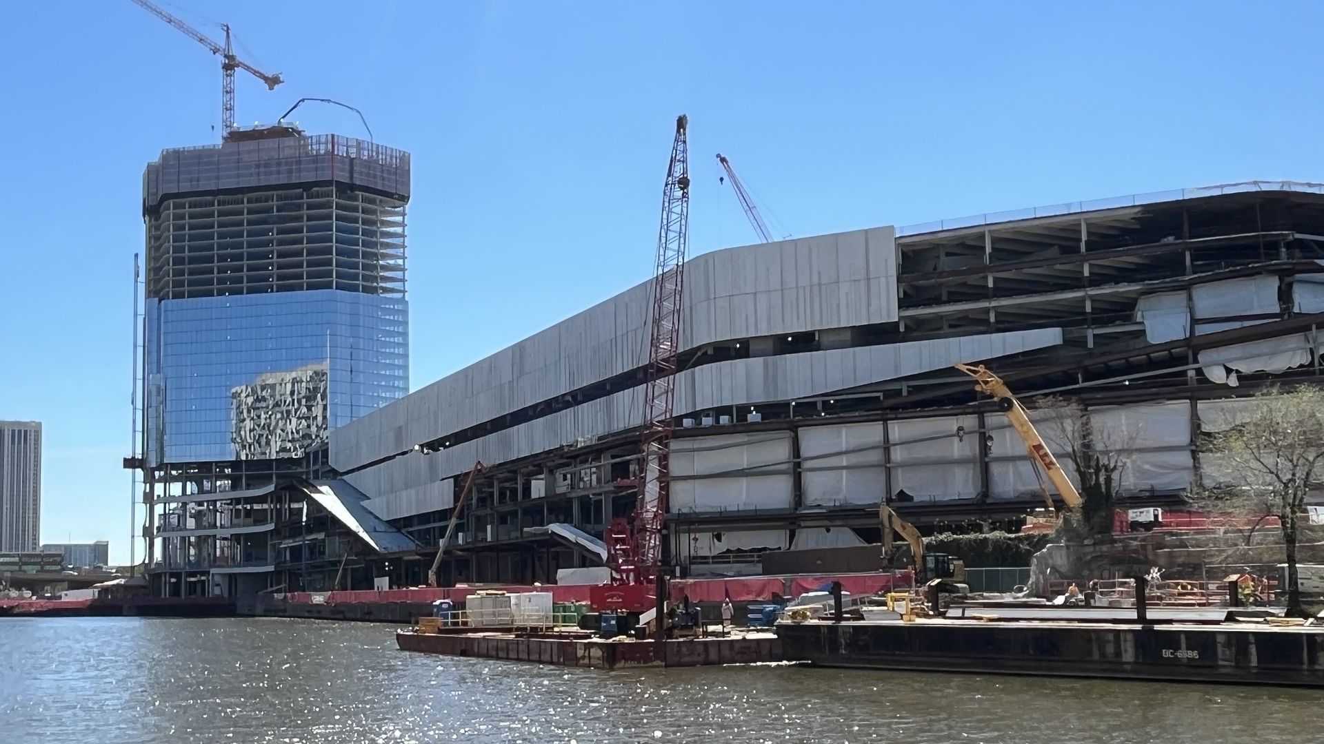 Construction site along a river: a tall glass-clad tower with a crane on top left, another crane near the center, and a curved, partially completed multi-level structure on the right beneath a blue sky.