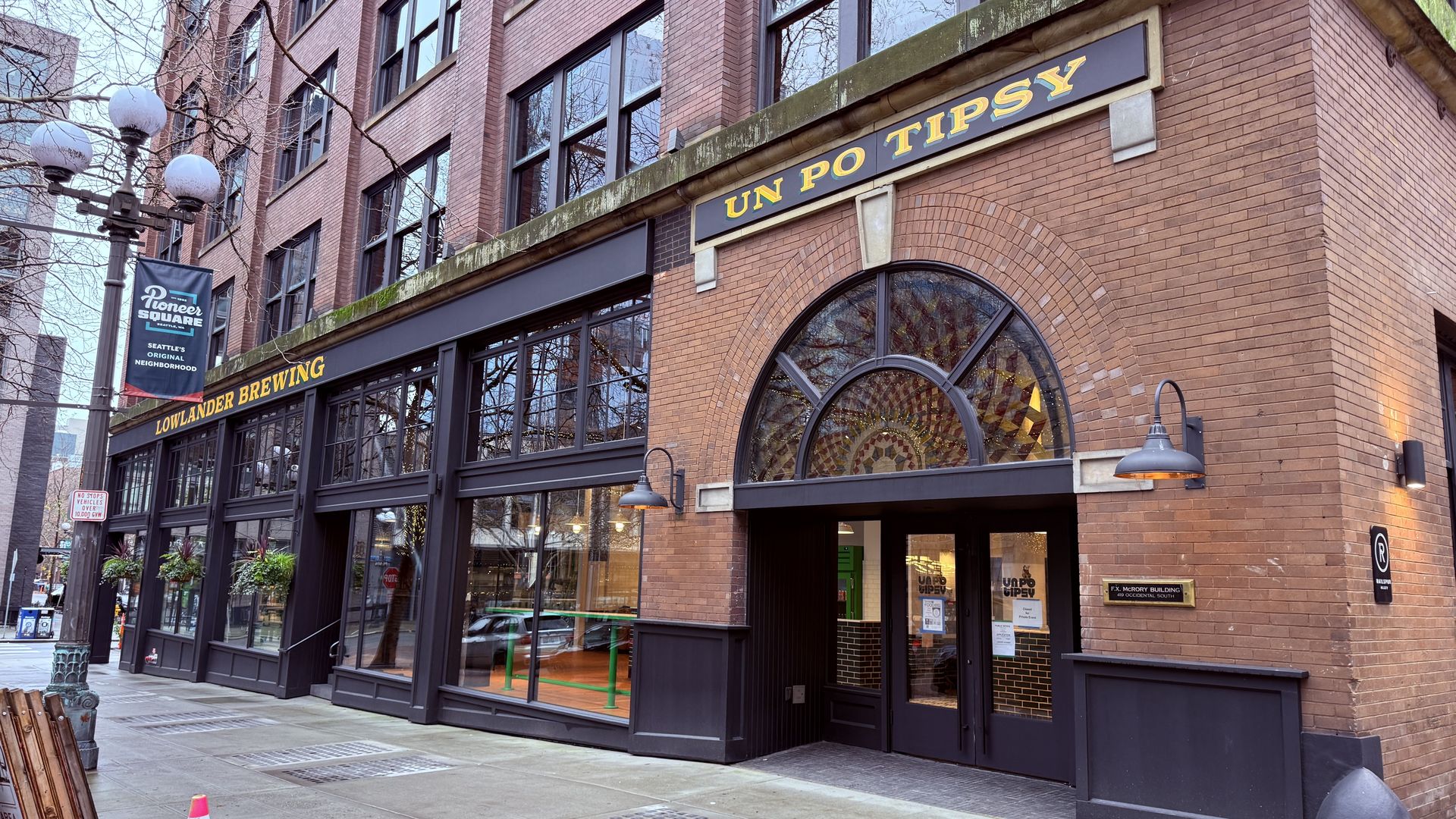 Exterior of a brick building on a city street with signs reading "LOWLANDER BREWING" and "UN PO TIPSY" above two black door entrances. A street lamp and traffic cone are visible.