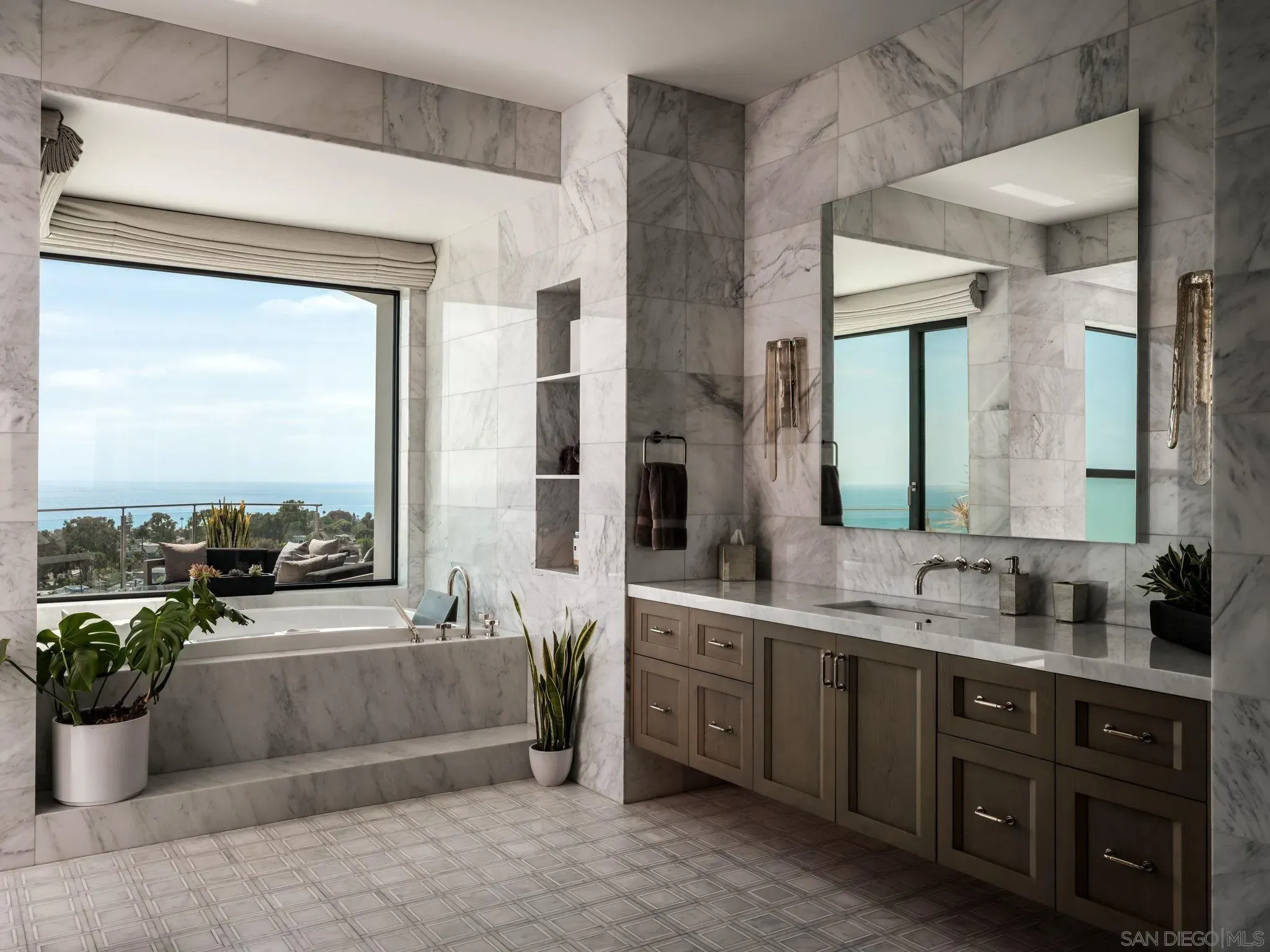 Luxurious marble bathroom with gray-white veined walls, large window overlooking blue sea, built-in tub, wood vanity with chrome faucets, green plants, and a wide mirror reflecting the space.