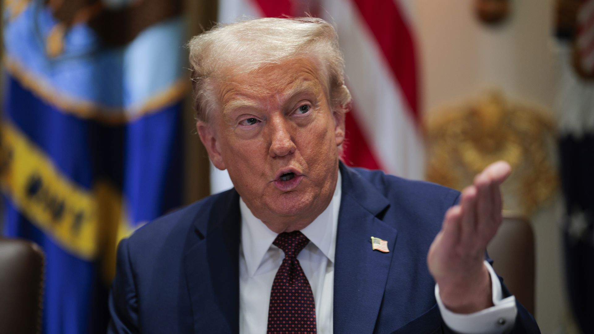 A man with light blond hair, President Trump, in a navy suit and red patterned tie gestures with his right hand, speaking with an expressive face. Blurred flags and gold decor are in the background.