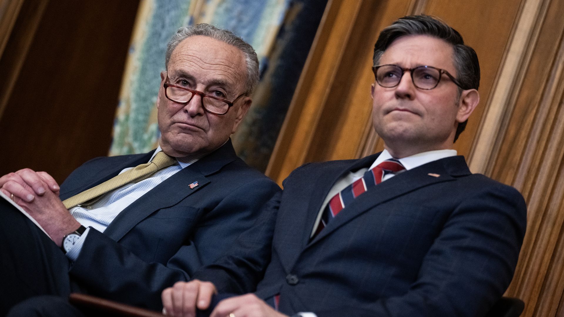Chuck Schumer, wearing a dark blue suit, light blue shirt and gold tie, and Mike Johnson, wearing a dark blue suit, light blue shirt, red-and-blue striped tie and glasses, sitting in front of a wood-paneled wall.