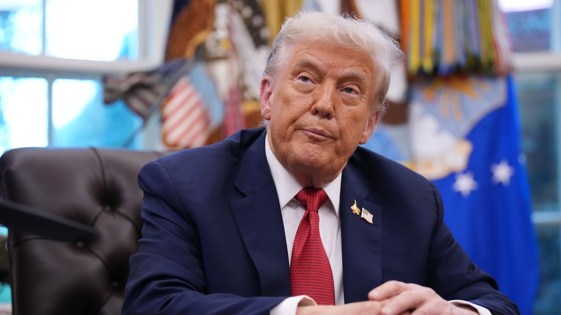 President Trump, wearing a navy jacket with a U.S. flag pin on it, white shirt and red tie, purses his lips as he sits with his hands clasp on a red leather chair at a desk in the Oval Office of the White House. 