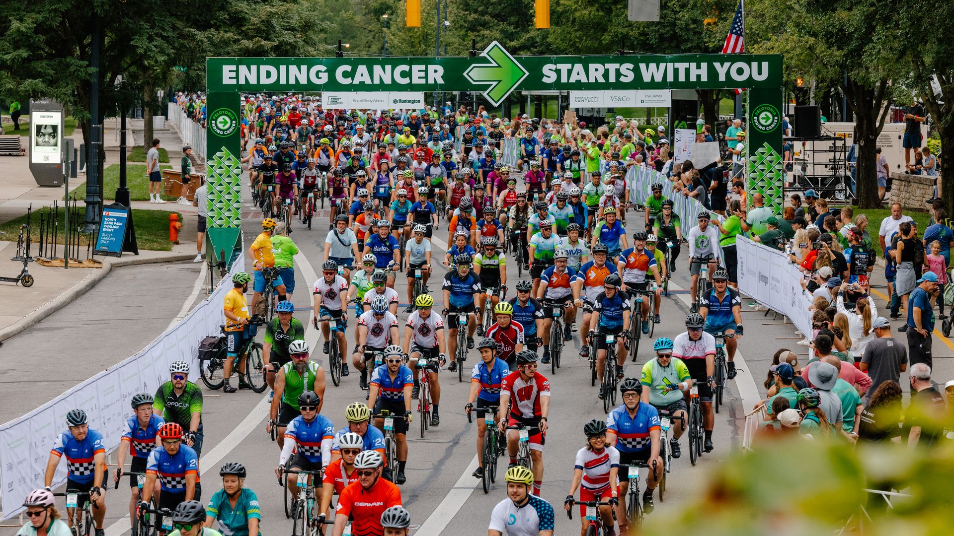 Hundreds of cyclists participating in a charity ride under a green banner that reads "ENDING CANCER STARTS WITH YOU," surrounded by cheering spectators on a tree-lined street.