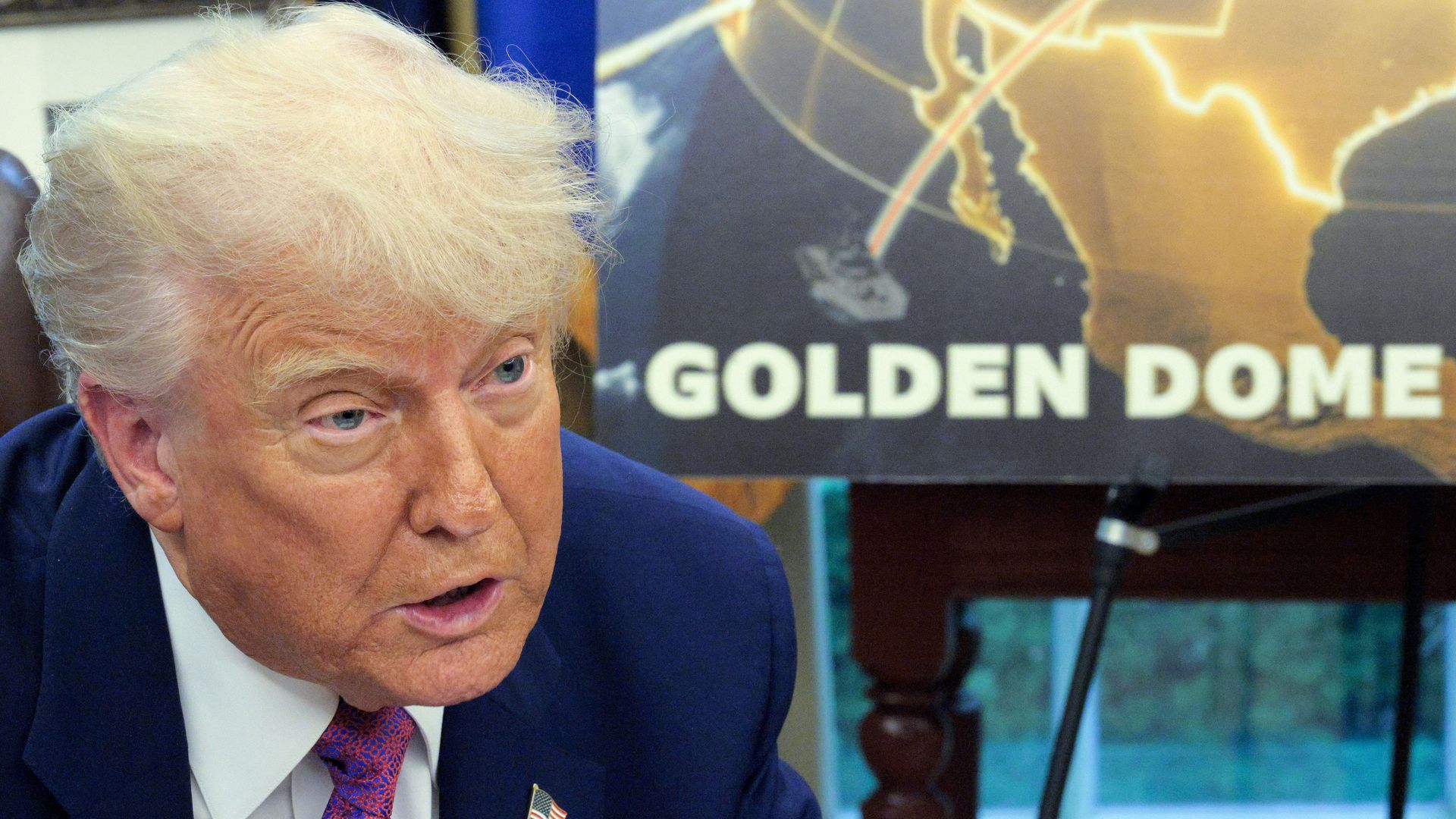 Close-up of a man with blond hair wearing a navy suit, white shirt, and red patterned tie with an American flag pin, speaking in front of a board showing part of a map and the text "GOLDEN DOME".