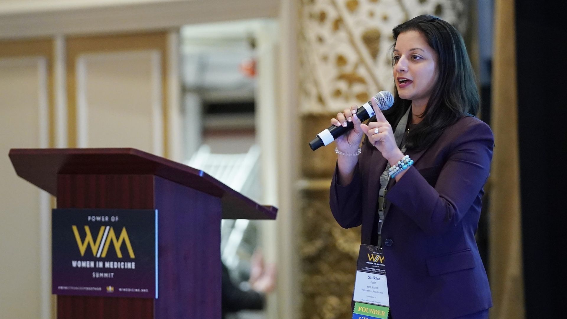 Female speaker in a purple blazer speaks into a handheld microphone at a wooden podium during a Women in Medicine conference; she wears a lanyard with multiple ribbons.