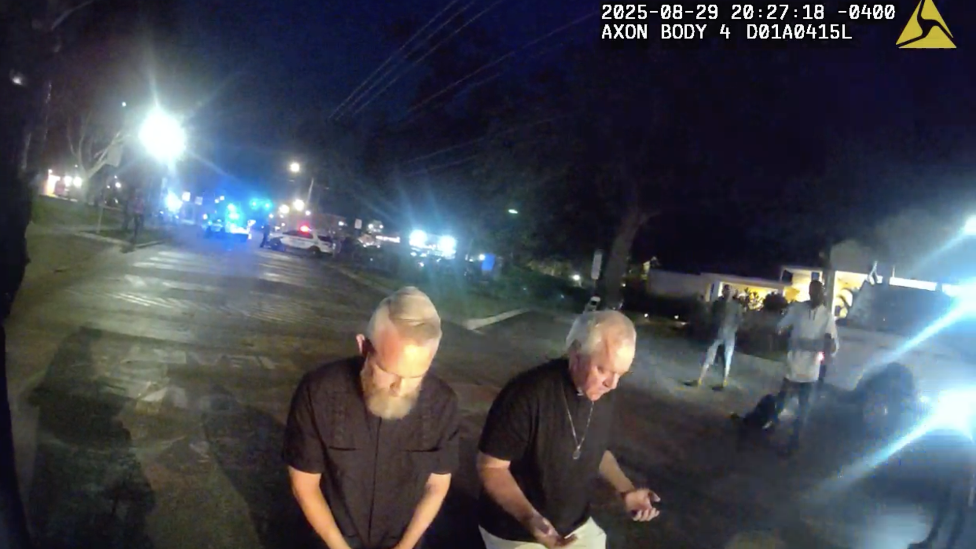 Two men in black shirts and clerical collars kneeling on the ground with their heads bowed. Surrounding them are several police cars with lights flashing.