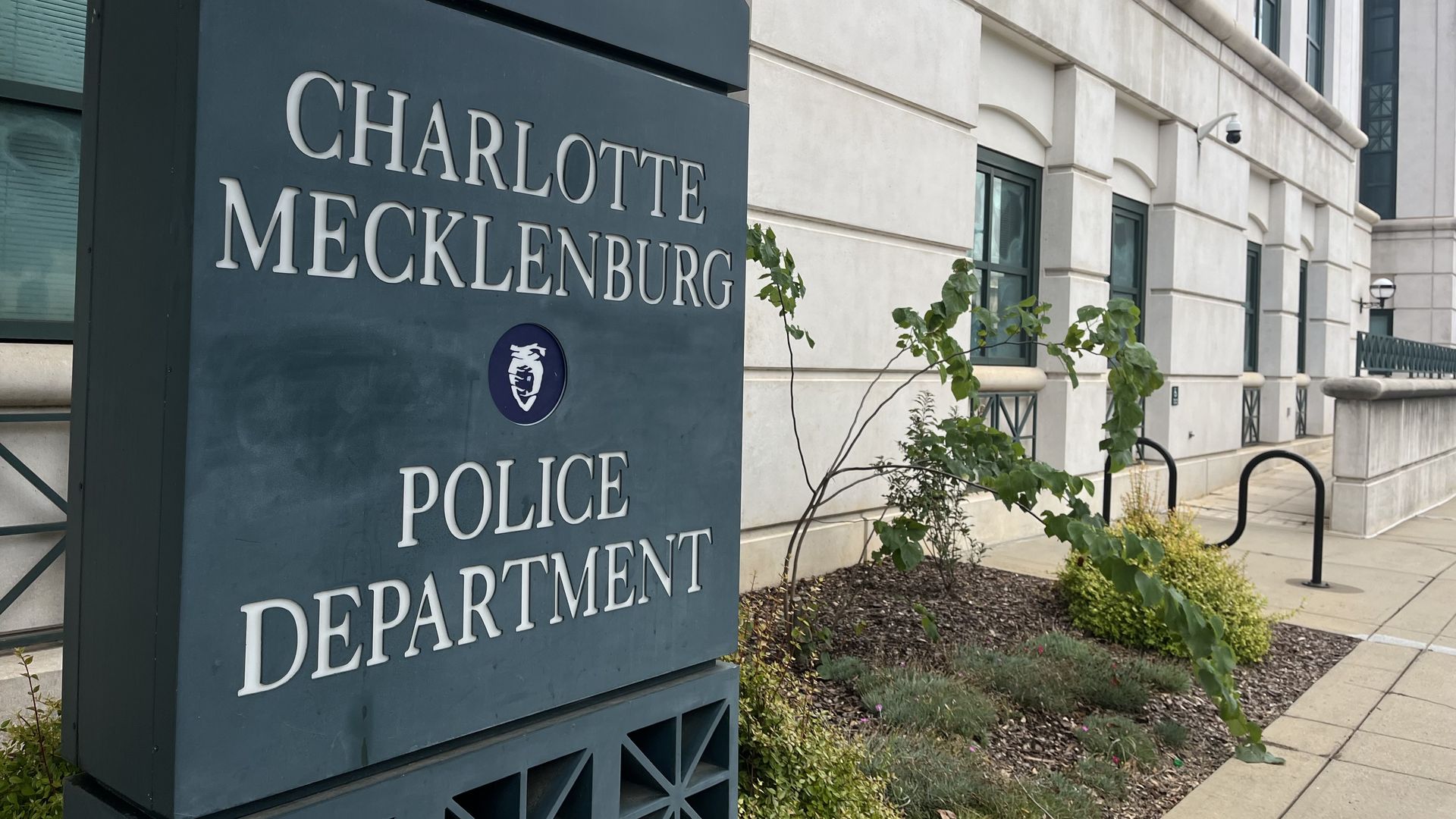 Exterior of the Charlotte Mecklenburg Police Department building with a blue sign displaying its name, beige walls, green windows, and some landscaping with plants and bushes.