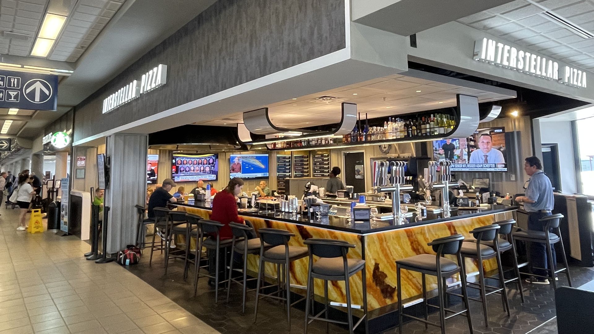 Bar-style pizza restaurant named Interstellar Pizza in a busy airport terminal, featuring yellow-orange stone bar counter, several customers seated, multiple TVs, and overhead liquor bottles.