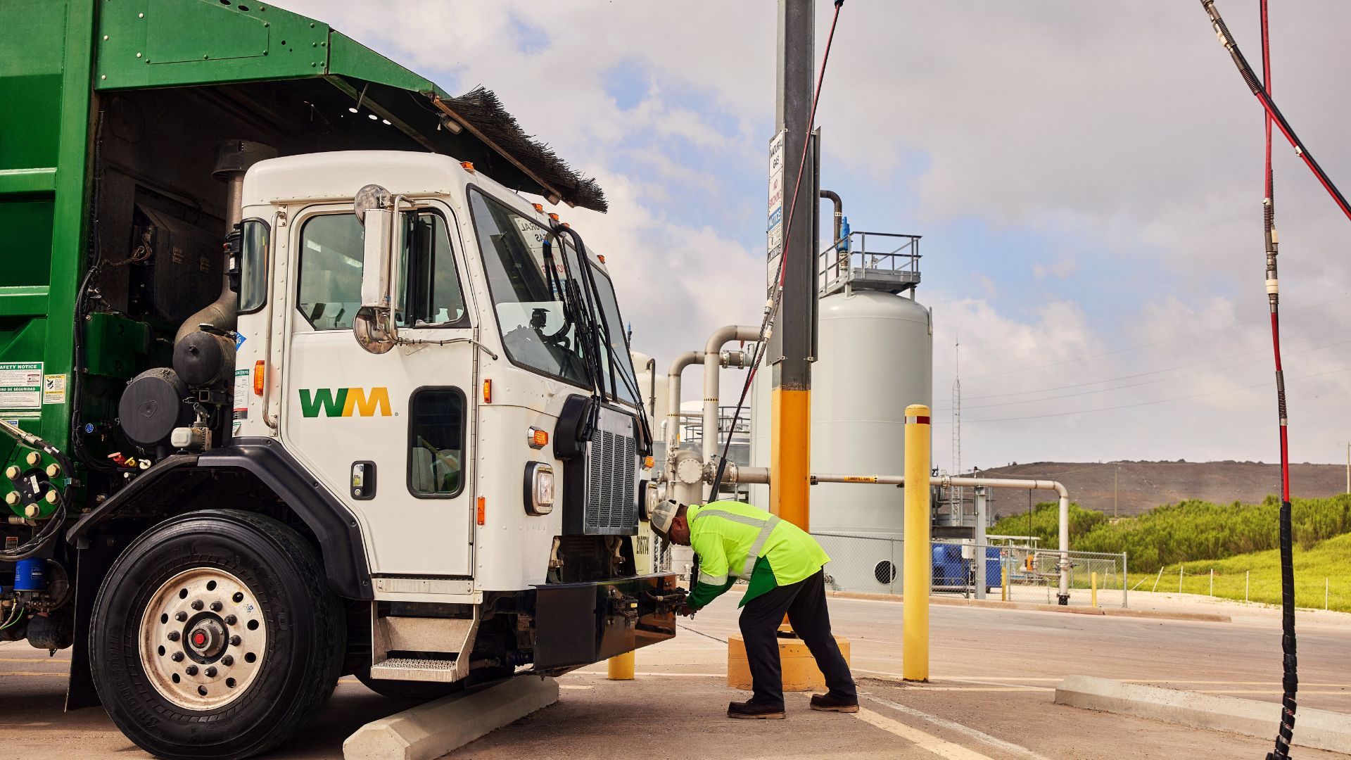 Worker in a neon yellow safety jacket inspects the front of a white and green Waste Management truck near industrial tanks under a partly cloudy sky.