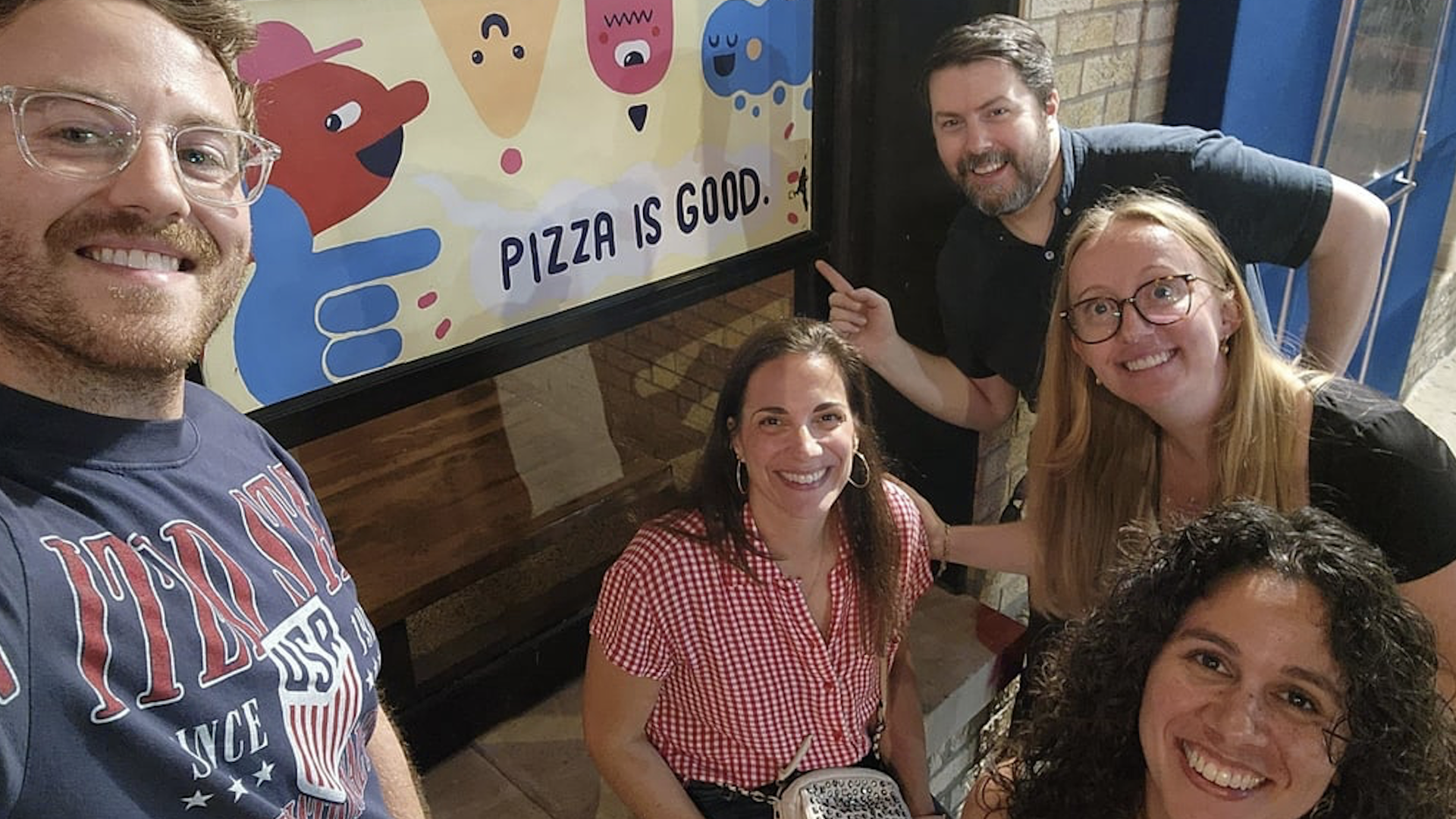 A group of friends taking a selfie in front of a storefront window with a colorful banner reading "PIZZA IS GOOD," featuring cartoon shapes and smiling faces against a brick wall.