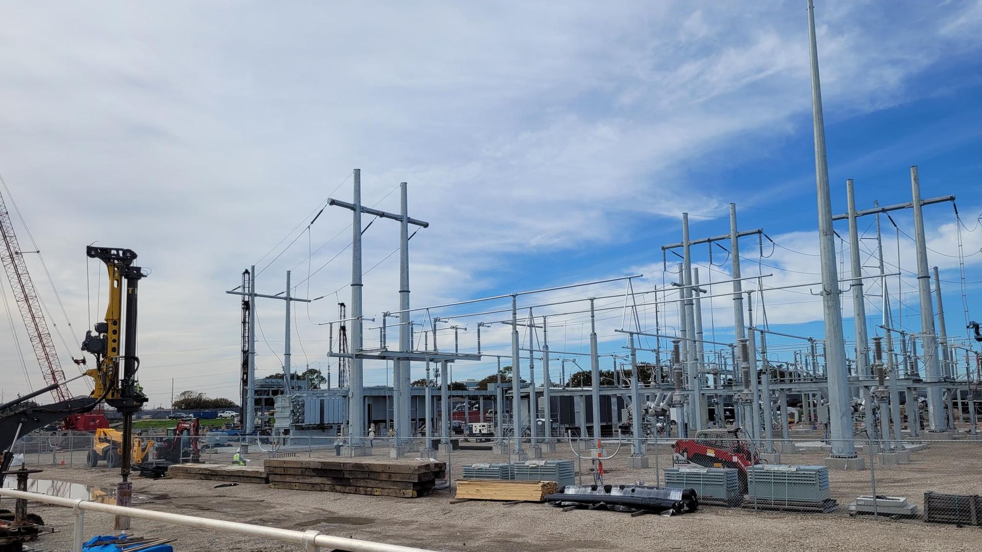 Construction site with gray steel electrical frames and poles, wires and scaffolding, a crane on the left, orange hoses on the ground, and a blue sky.