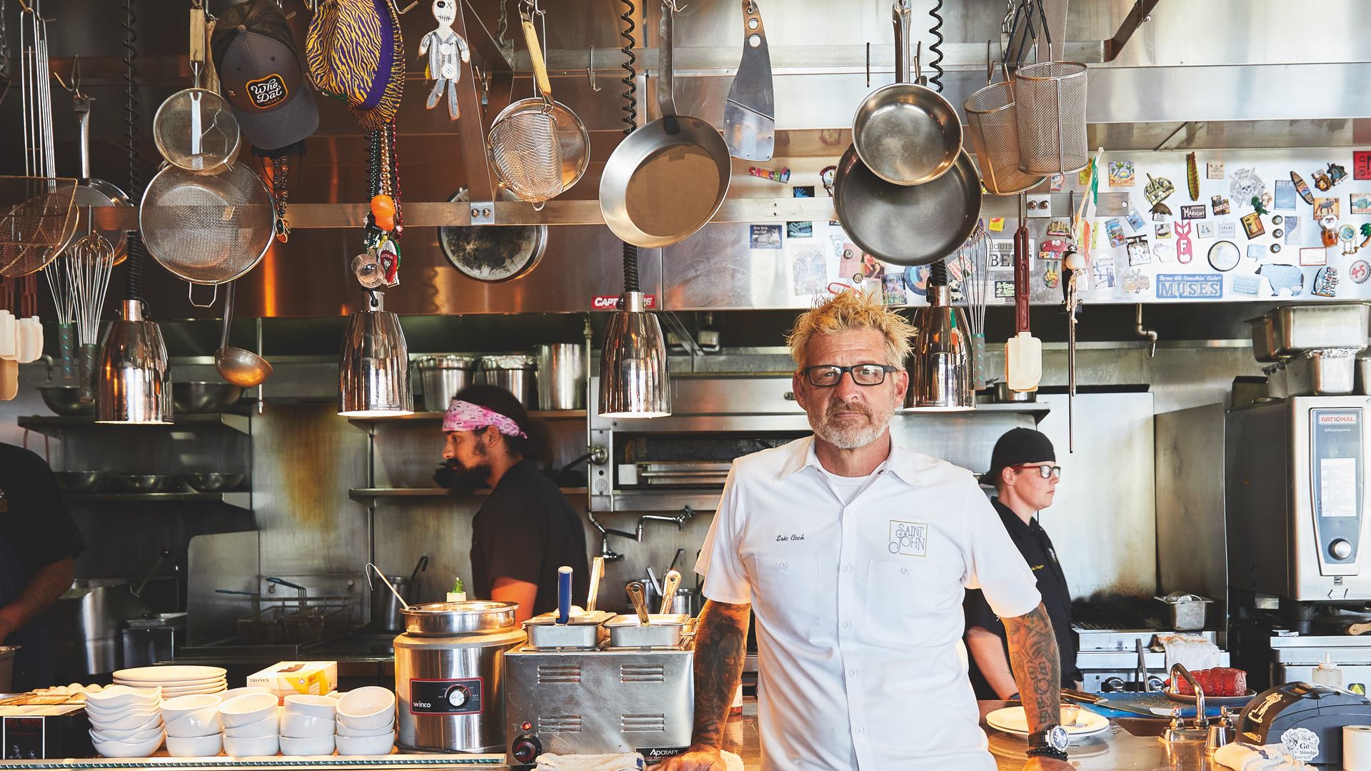 Chef Eric Cook poses for a photo in a busy kitchen. He looks directly at the camera as pots hang overhead.