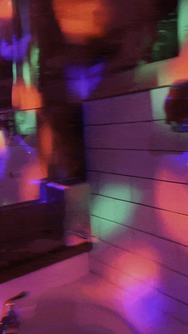 Dim interior bathed in neon purple, blue, green, and red lights reflecting off wood-paneled walls and tiled surfaces; blurry silhouette on the right, bottle on the left.