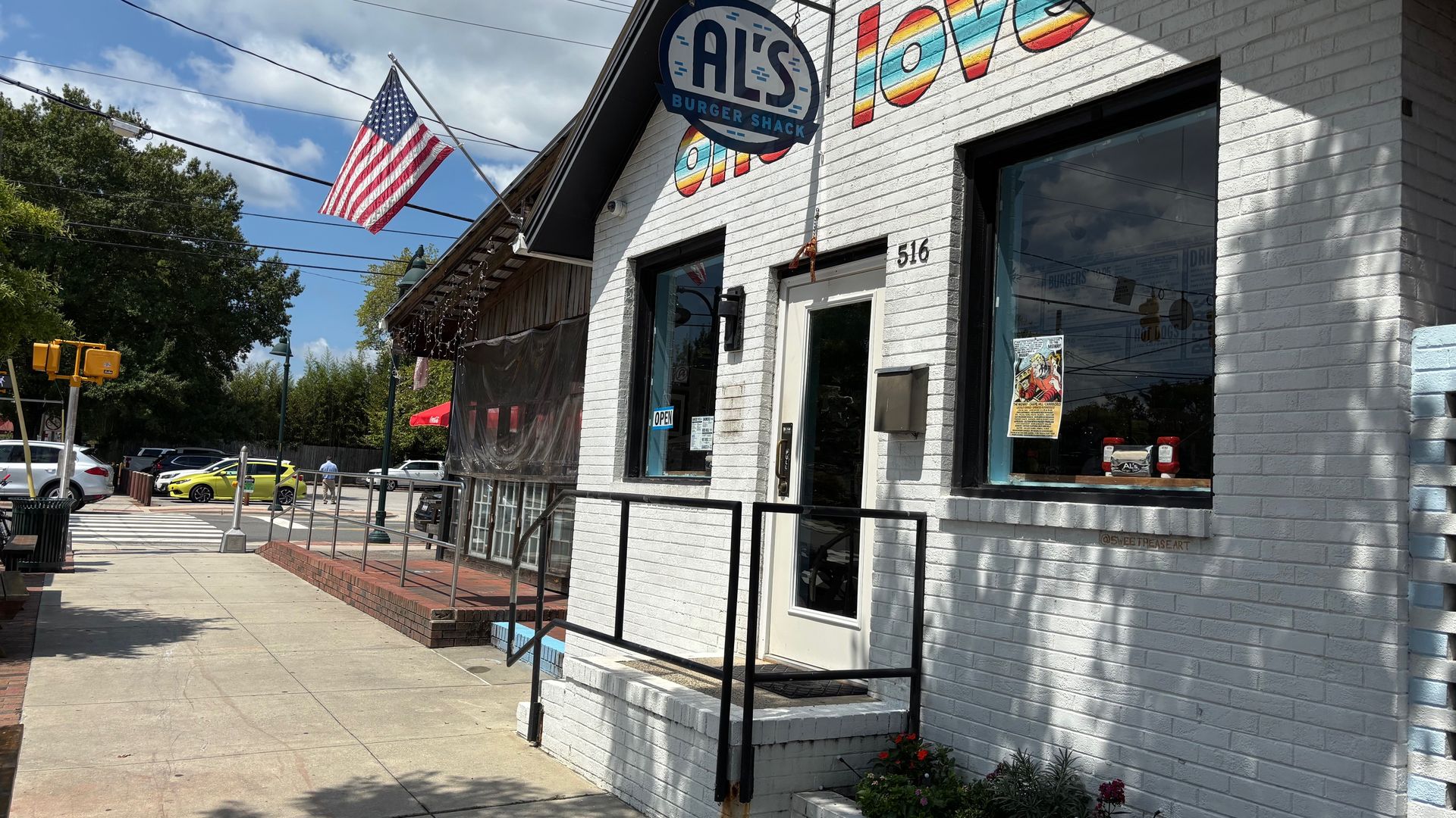 White brick building with a round blue sign reading "Al's Burger Shack" and colorful rainbow letters spelling "love". American flag on a pole, cars, sidewalk, and flowers in front.