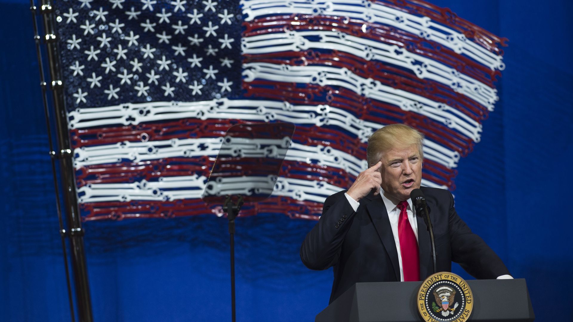 Trump in front of an American flag made of tools before he signed the Buy American, Hire American executive order. HIs index finger is pointing toward his head and he's speaking at a podium. 