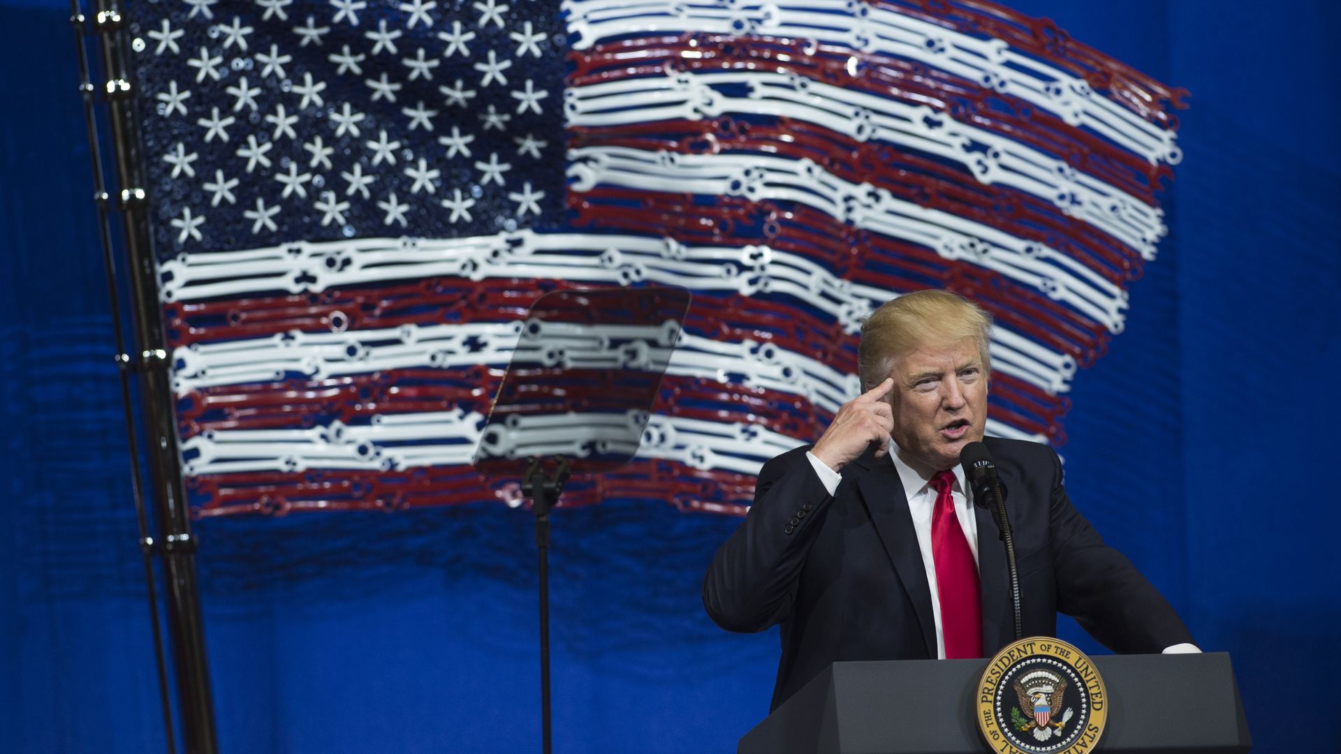Trump in front of an American flag made of tools before he signed the Buy American, Hire American executive order. HIs index finger is pointing toward his head and he's speaking at a podium.