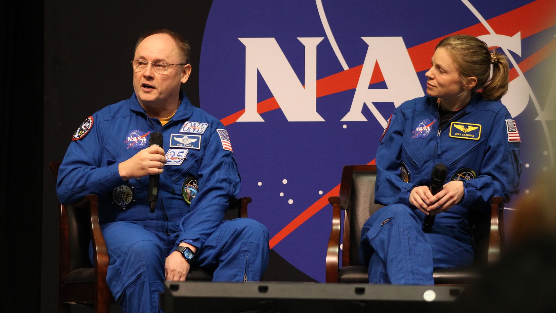 Two astronauts in blue NASA jumpsuits sit on stage in front of a large NASA backdrop. The man on the left speaks into a microphone; the woman on the right listens, both holding mics.