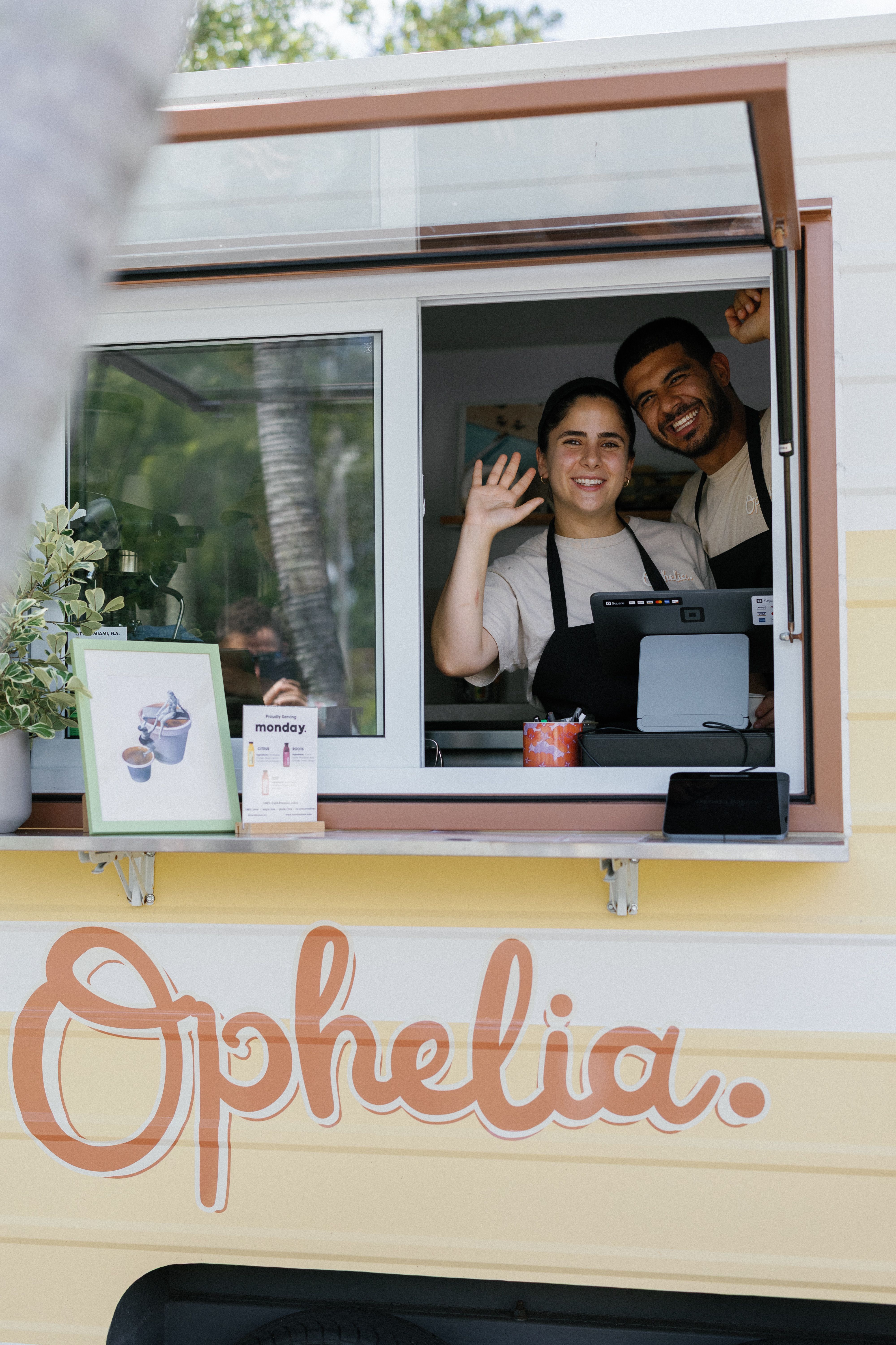 Ana De Sa Martins (left) and Juan Vieira (right) in light gray shirts and black aprons stand in a yellow food truck window, waving at the camera. A menu, plant, and sign sit on the counter with large orange script on the truck.
