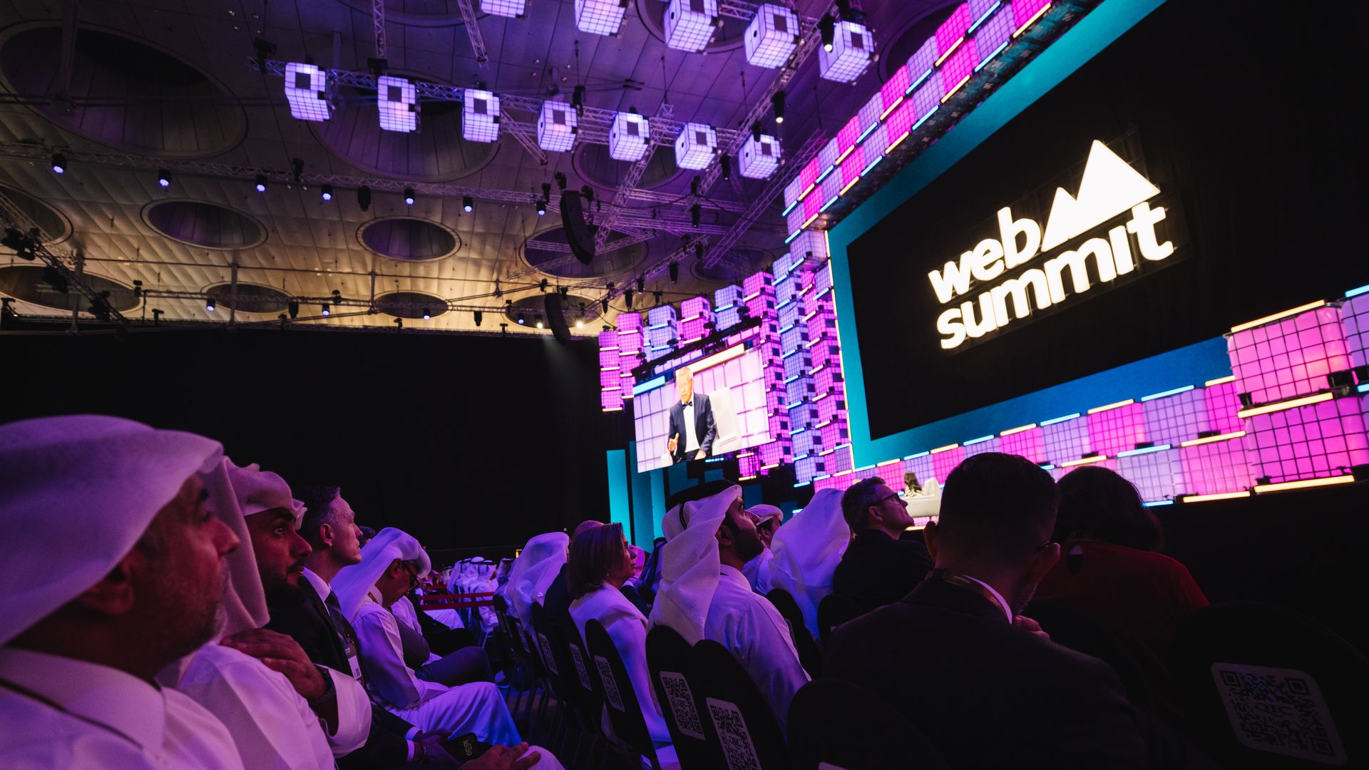 Audience in traditional Middle Eastern attire and business suits watching a presentation at the Web Summit conference, with purple and blue lighting and a large screen displaying the logo.