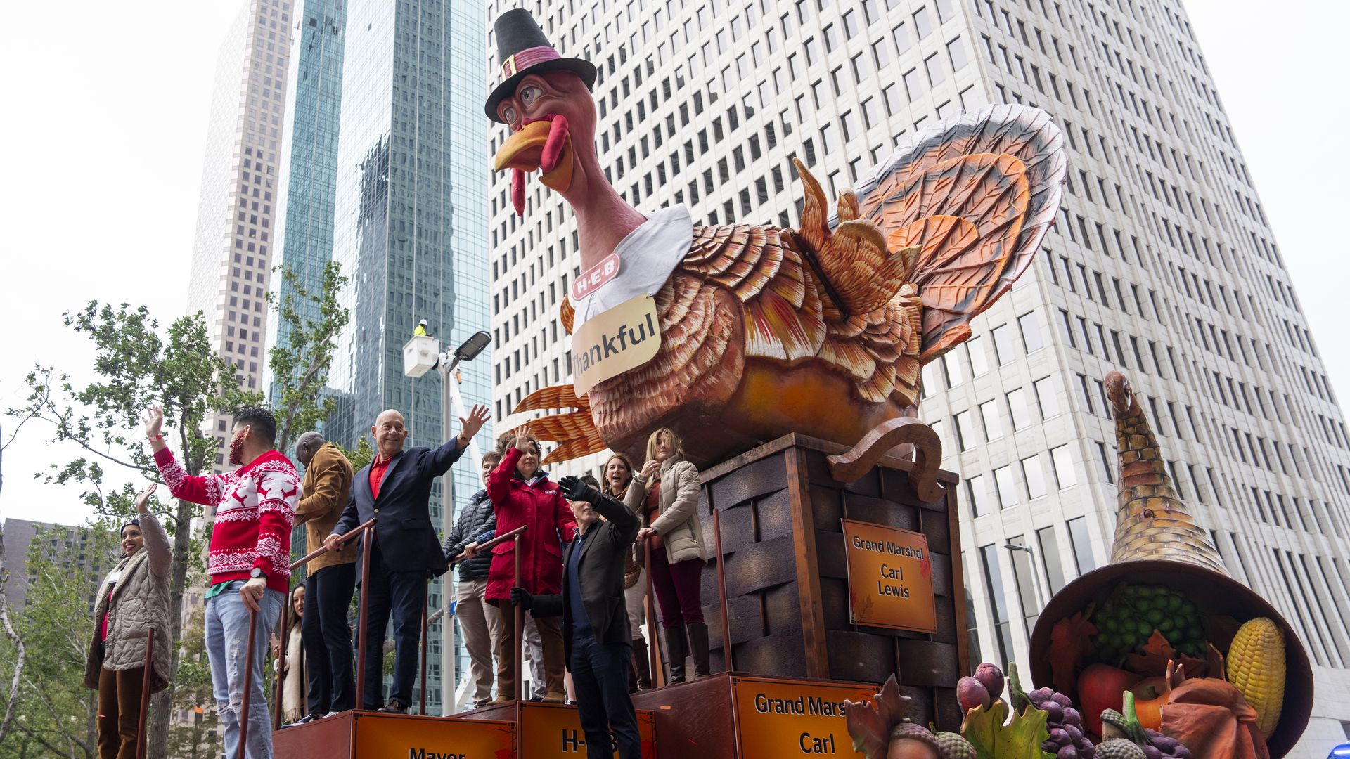Parade float with a large turkey wearing a pilgrim hat and "thankful" sign, surrounded by people waving. Skyscrapers and a cornucopia with fruits and vegetables nearby in the city.