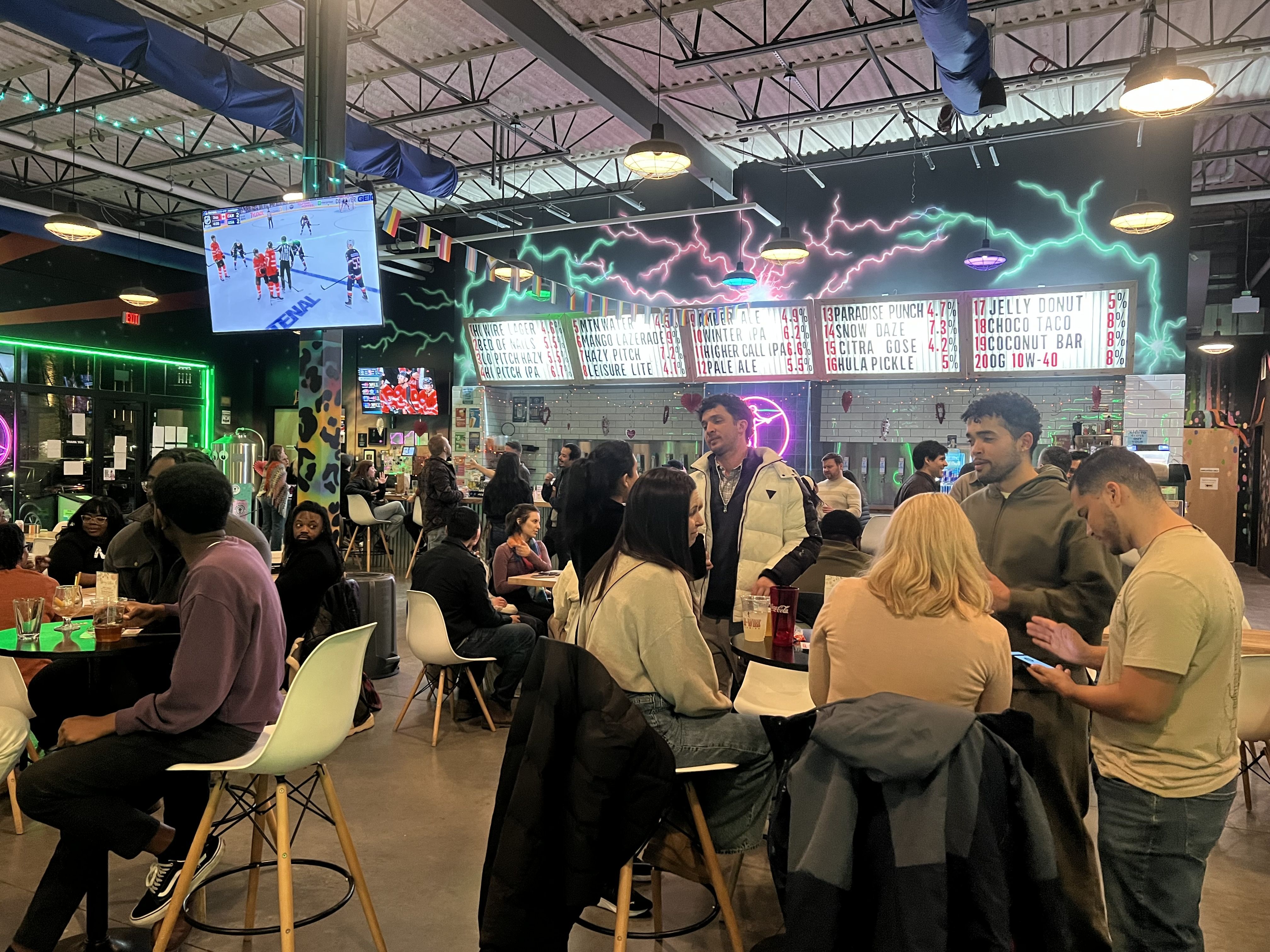 Crowded bar with neon signs, menu boards, and TVs showing a hockey game, featuring people socializing and sitting around tables in a vibrant, colorful indoor setting.