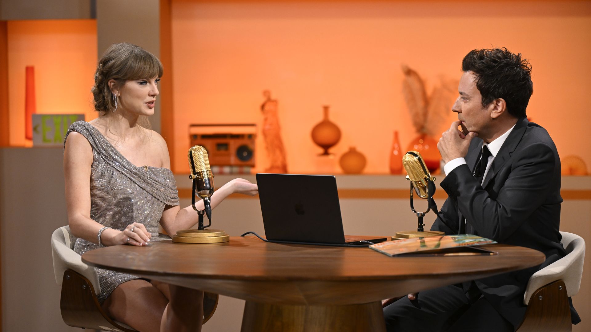 Singer-songwriter Taylor Swift, wearing a sparkly one-shoulder dress, and host Jimmy Fallon, in a black suit, discuss her latest album "The Life of a Showgirl" around a wooden table with an orange lit backdrop.