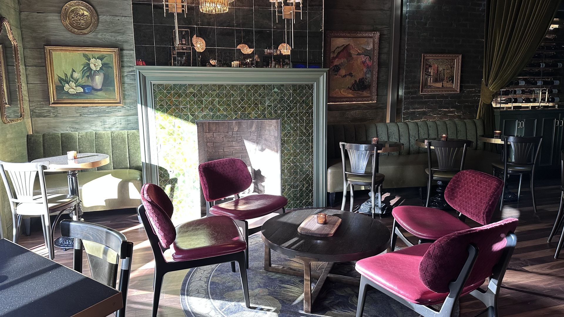 Cozy lounge with green padded benches, dark wood tables, and four red cushioned chairs around a round wooden coffee table; fireplace with green tiles and framed paintings on walls.