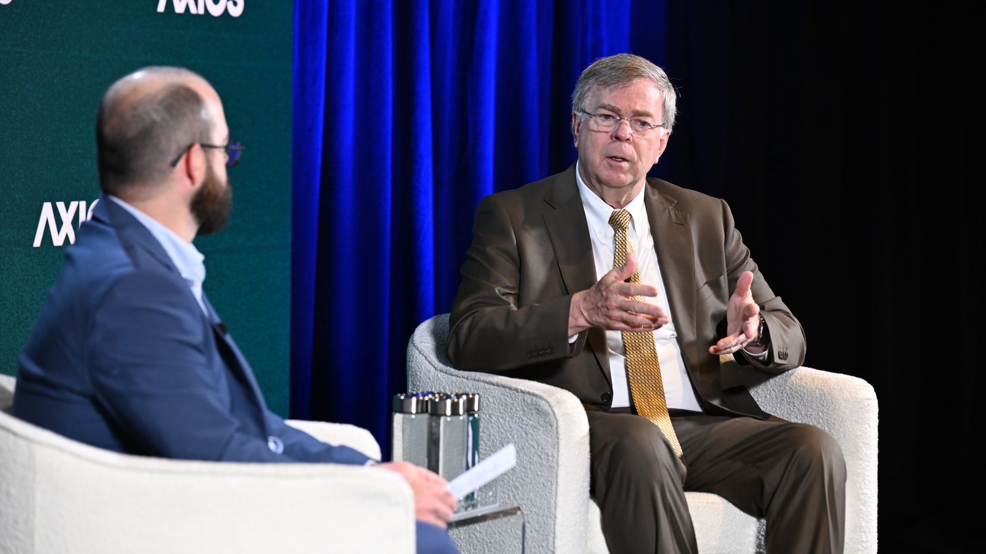 Two men in a panel discussion on a stage. One in a brown suit and yellow tie speaks with hands; the other in a blue suit listens. Blue curtains and a green Axios backdrop behind them.