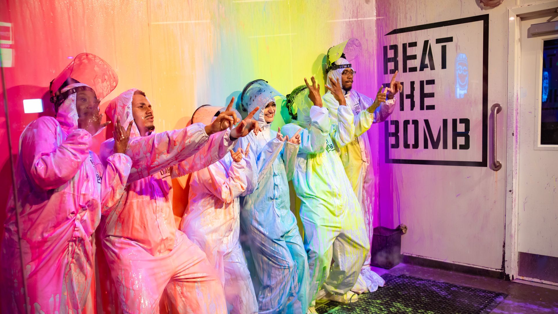 Several people in hazmat suits drenched in various paint colors next to a wall reading BEAT THE BOMB.