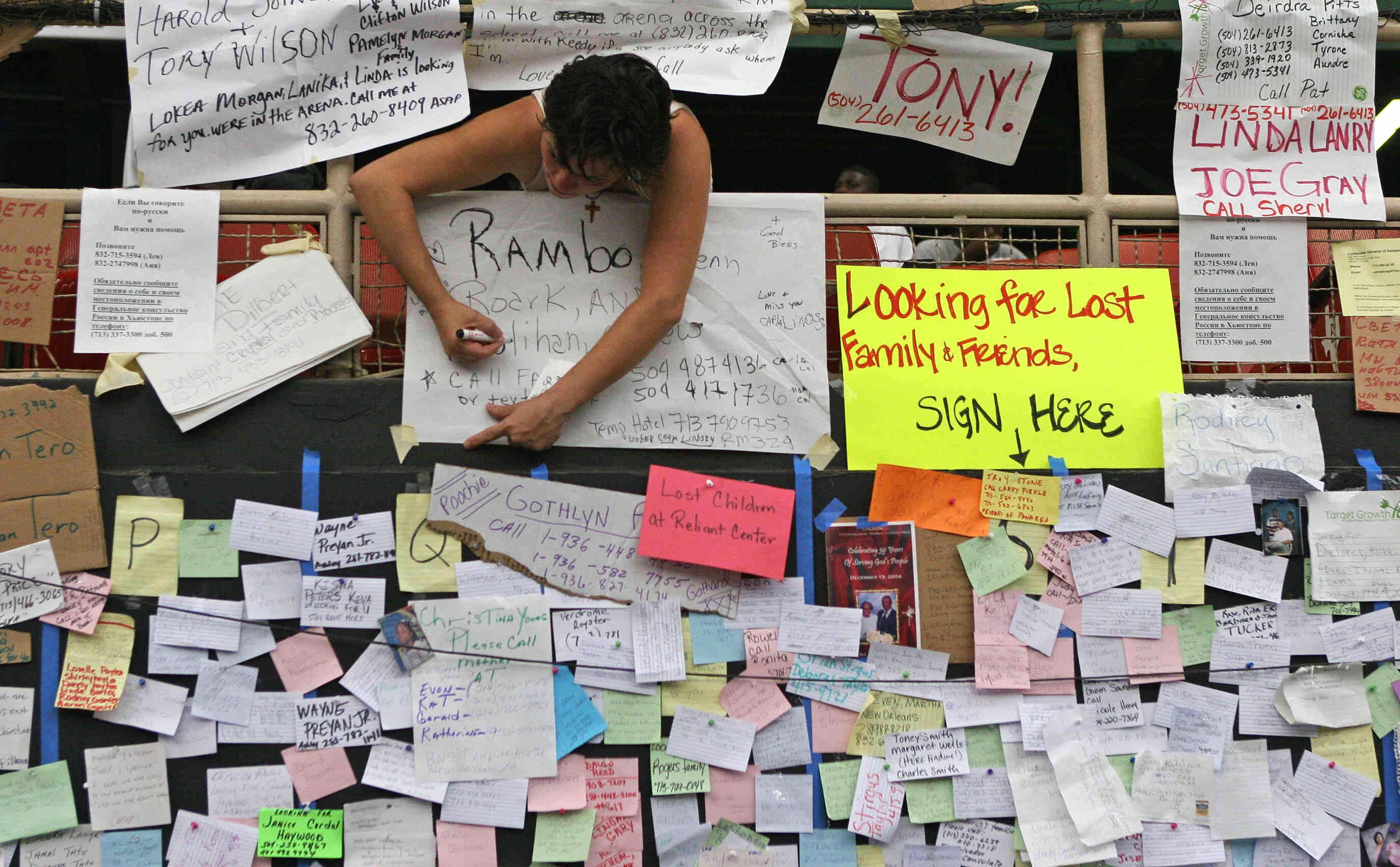 A person writes on a large poster among numerous handwritten notes and signs on a wall, including a bright yellow one reading "Looking for Lost Family & Friends, SIGN HERE."