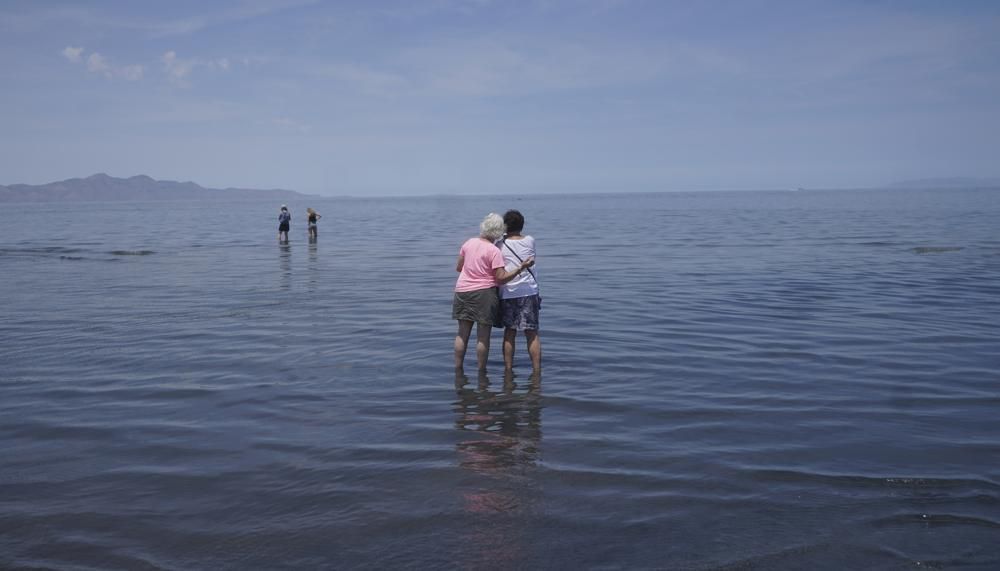 Two people with their arms around each other stand in shallow water and look towards the horizon of Utah's Great Salt Lake in June.