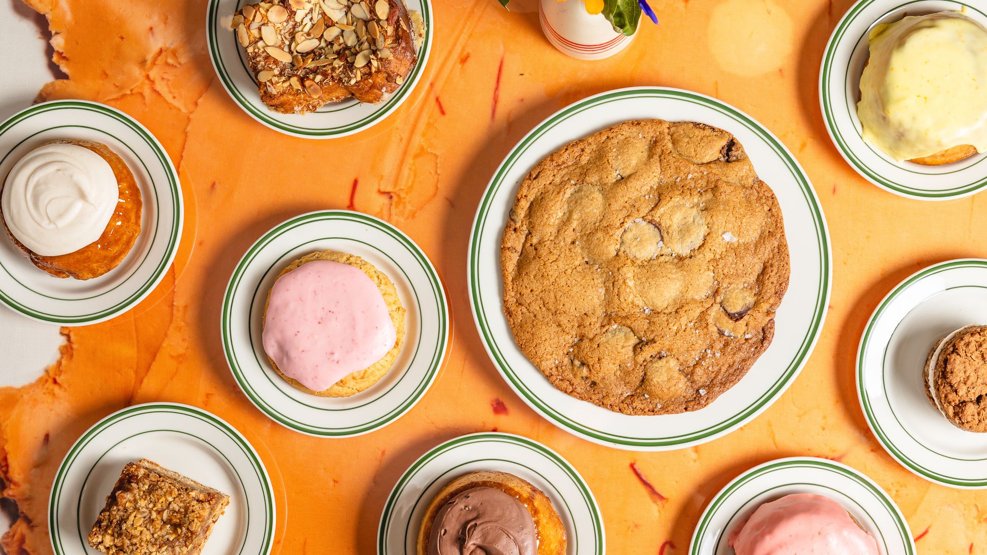 Top-down view of an orange tablecloth with green-rimmed plates surrounding a large oatmeal cookie, almond-topped pastry, pink and chocolate frosted donuts, a cream-topped pastry, and a vase of flowers.