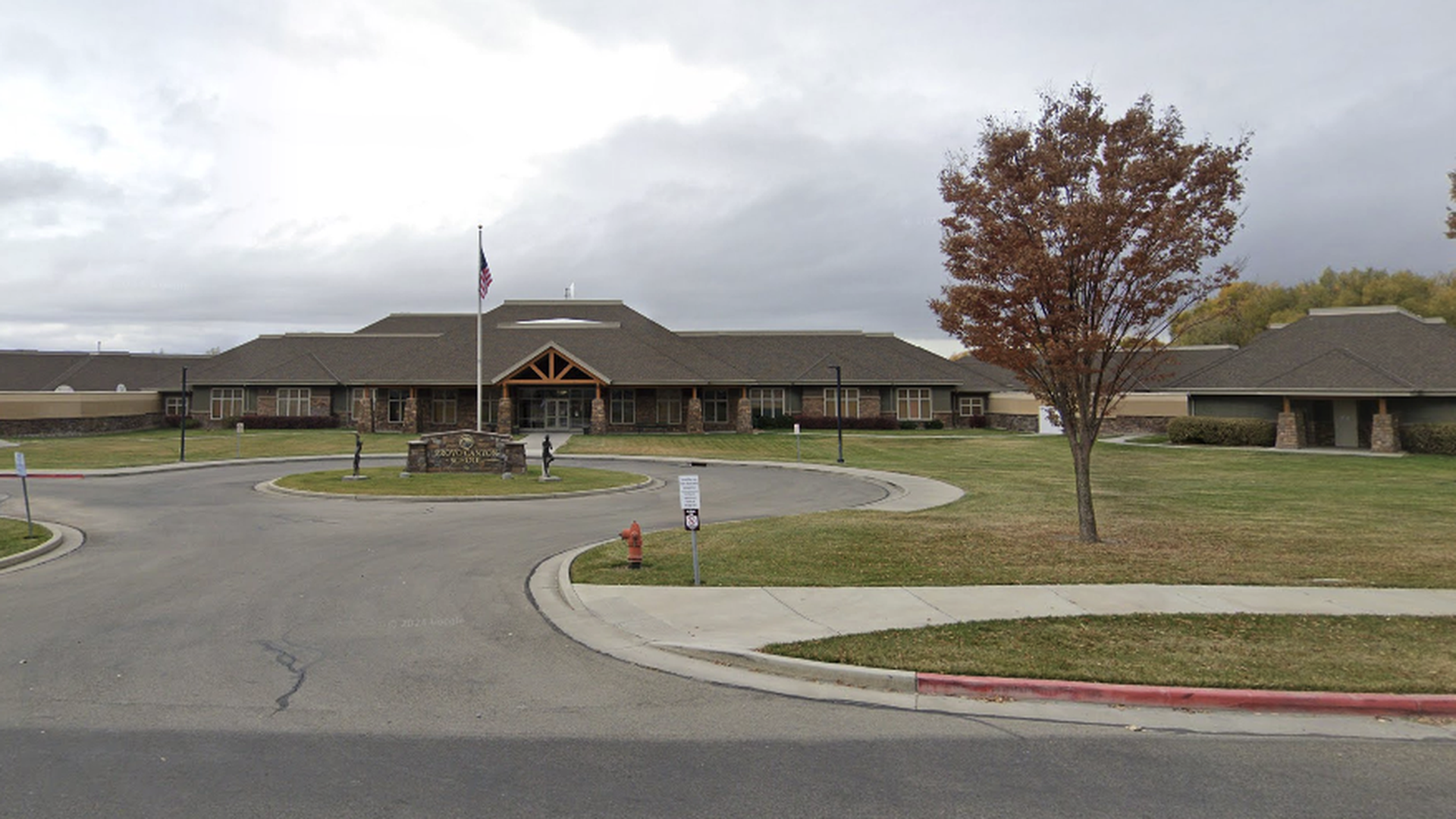 Single-story brick building with a peaked wooden entrance, set behind a circular driveway. An American flag flies near a stone sign; a brown tree with orange leaves stands to the right under a cloudy sky.