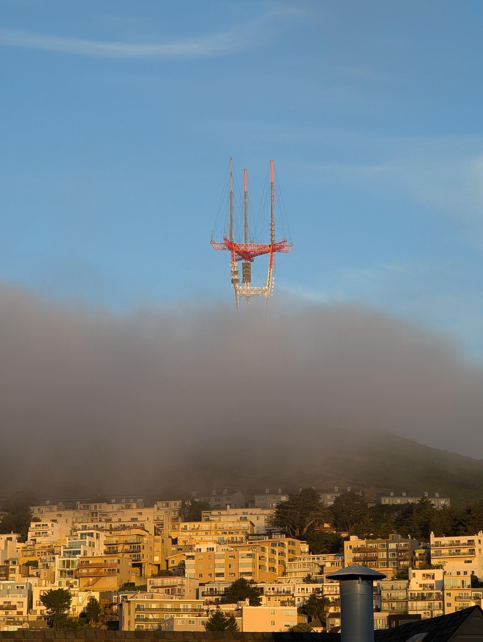 Tall red-and-white radio tower with guy wires rises above a fog-shrouded city of beige apartment buildings, under a clear blue sky.