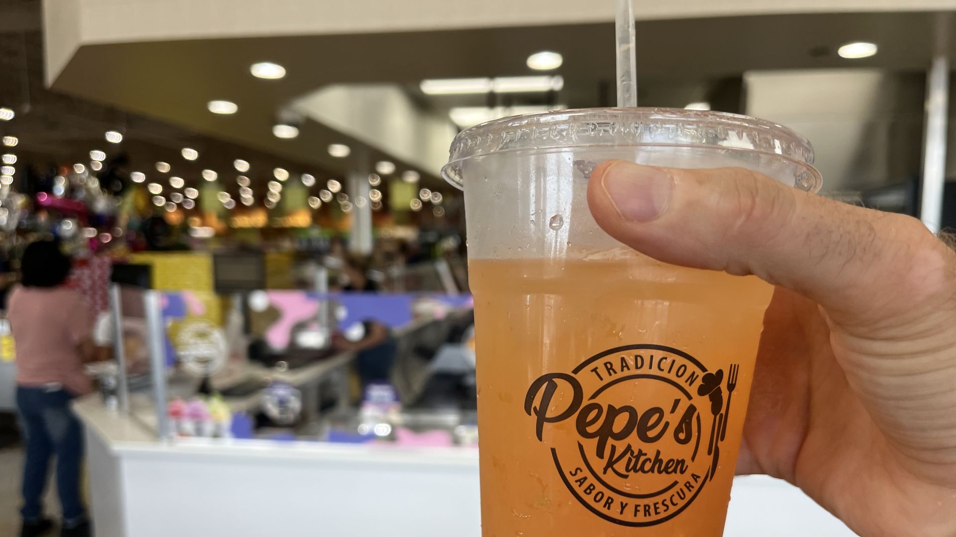 Close-up of a hand holding an orange beverage in a clear plastic cup with a lid; logo on the cup is visible. The background shows a blurry store interior with bright lights.