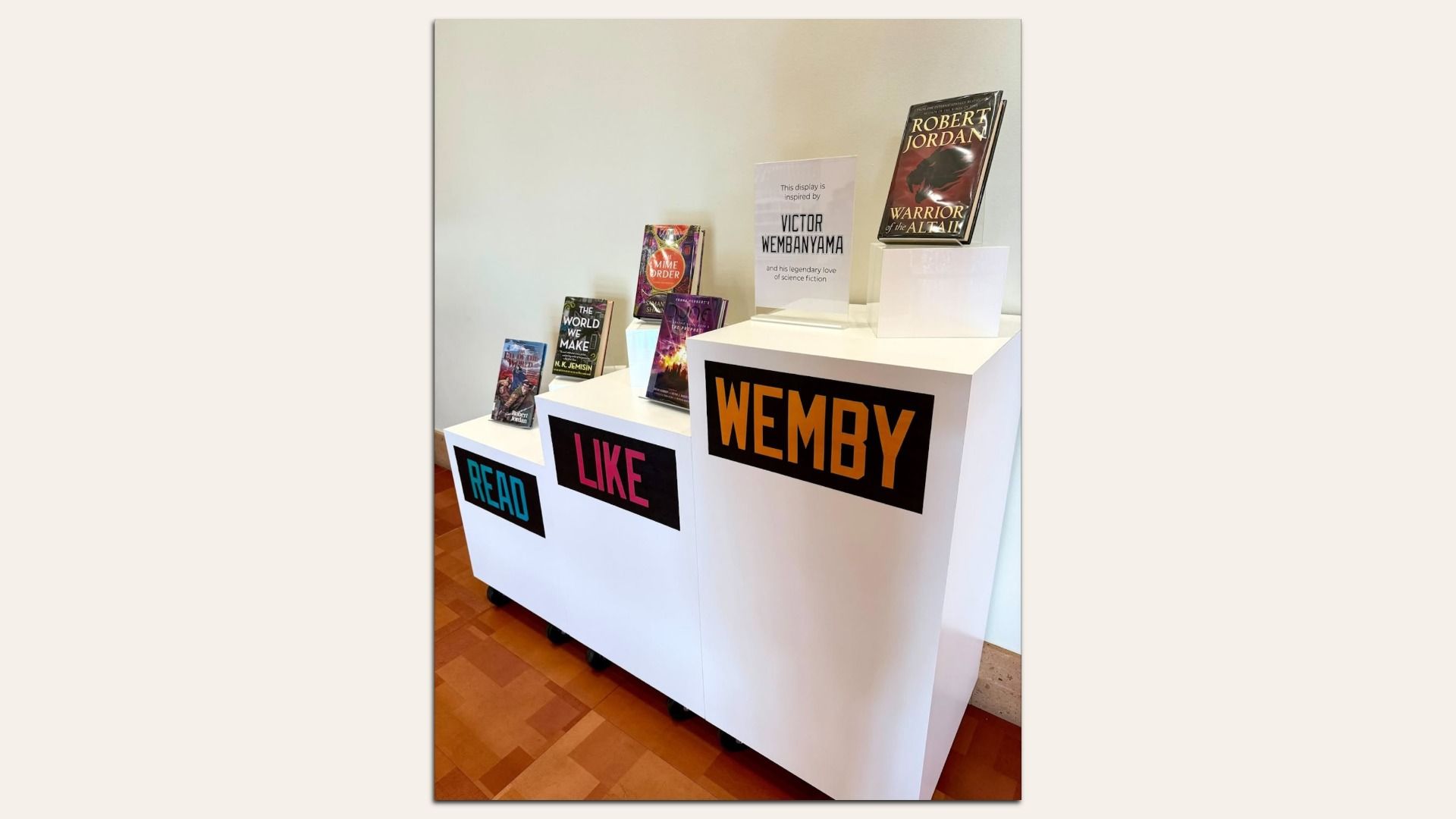 Exhibit with white pedestals labeled READ, LIKE, and WEMBY; small book covers displayed; a sign references Victor Wembanyama and Robert Jordan's "Warrior of the Altaii".