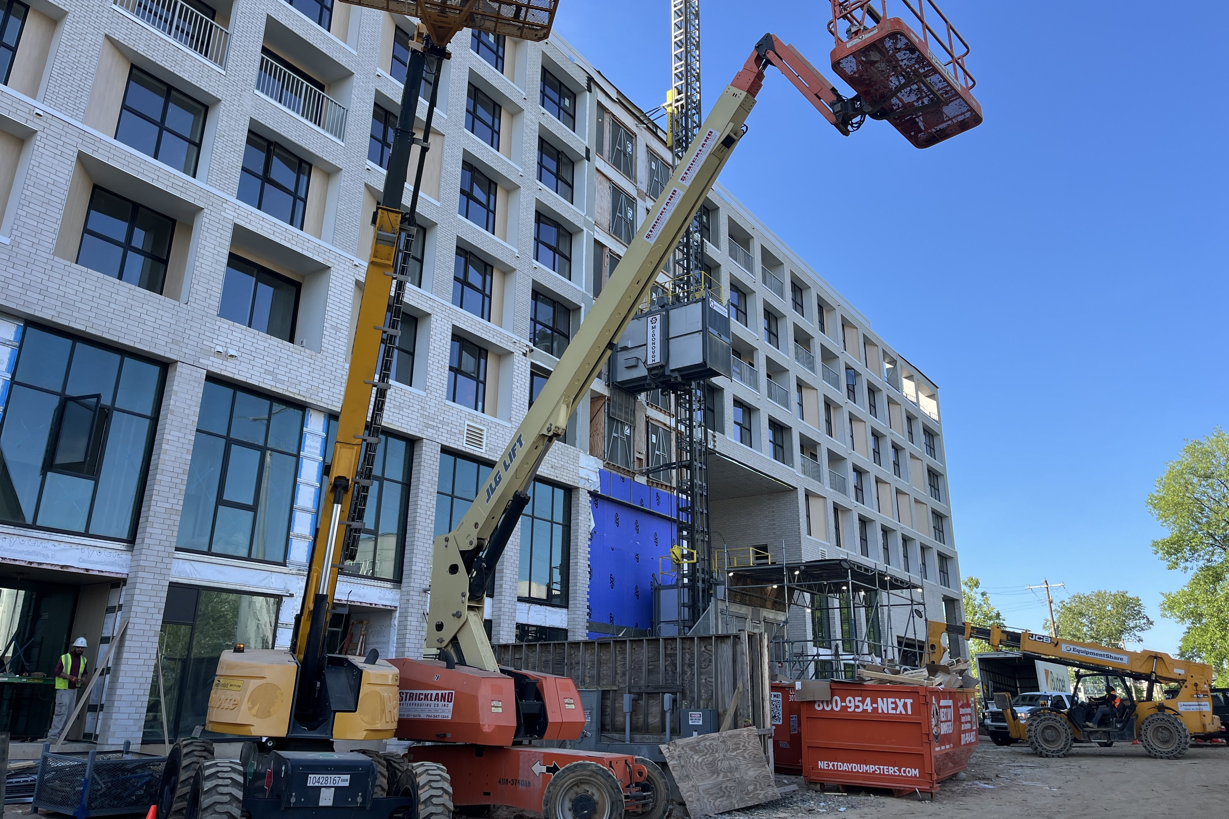 Construction site with a white brick, multi-story building under construction. Cranes and a lifting platform rise above scaffolding, while workers in safety vests work against a clear blue sky.