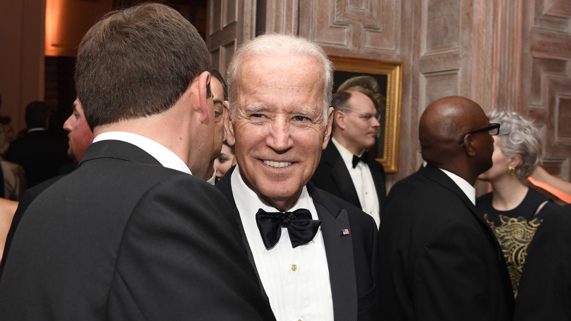 Joe Biden in a black tie and tuxedo at a party smiling with other guests around him