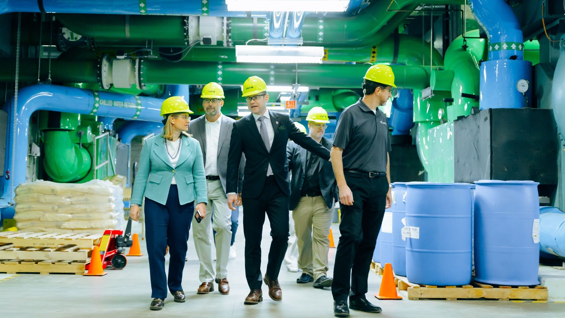 Five people wearing yellow hard hats touring an industrial facility with green and blue pipes overhead and blue barrels on pallets.