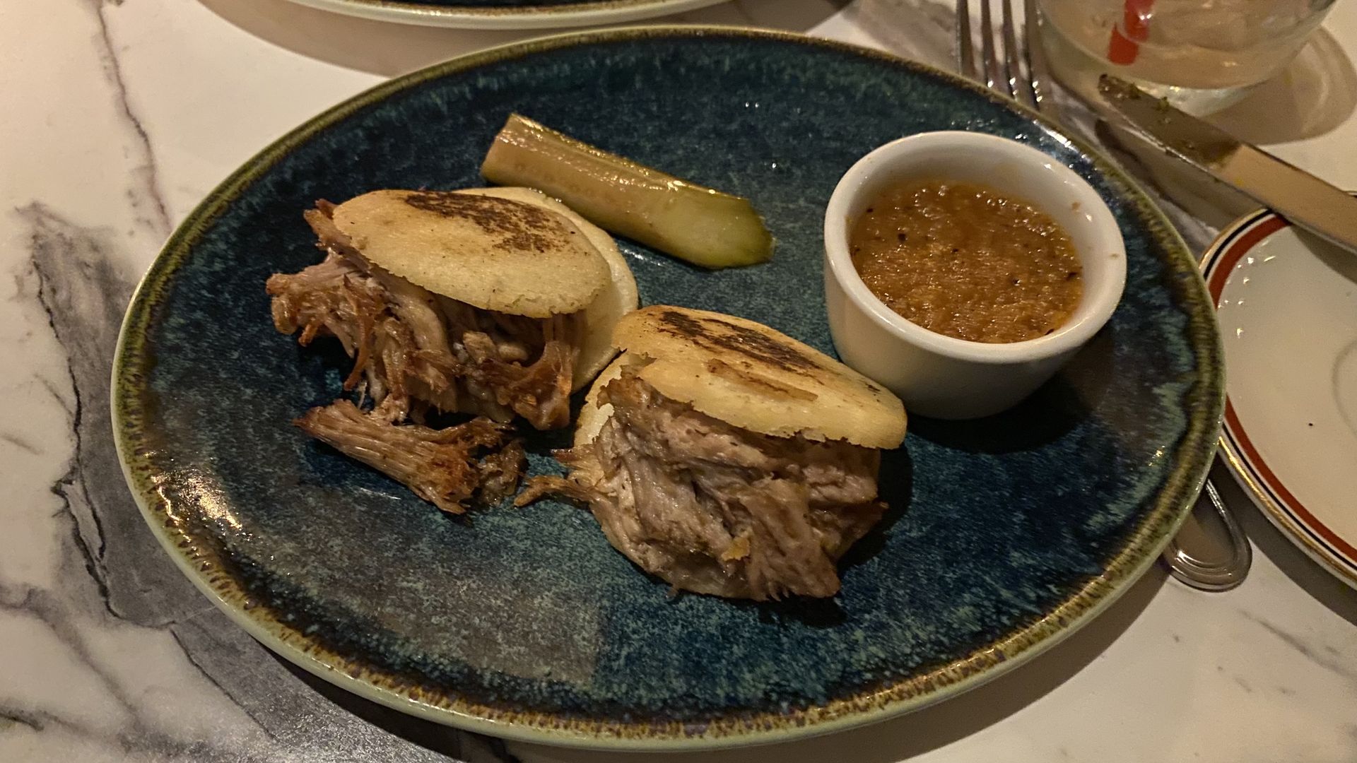Two small arepas filled with pulled pork on a plate next a small pickle spear and a cup of red sauce.