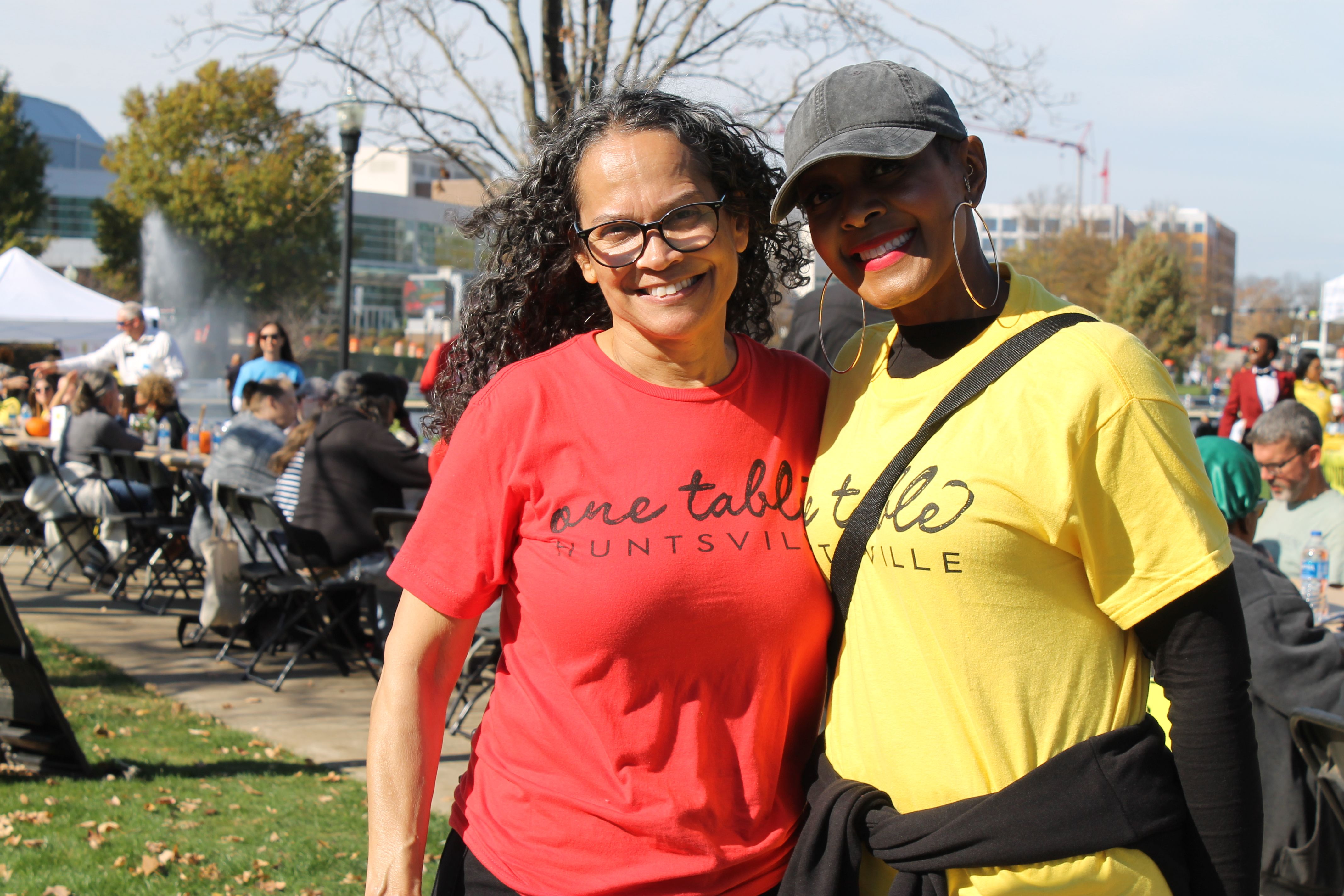 Two smiling women outdoors at a daytime event, one wearing a red "one table HUNTSVILLE" T-shirt and glasses, the other in a yellow T-shirt, black cap, and large hoop earrings.