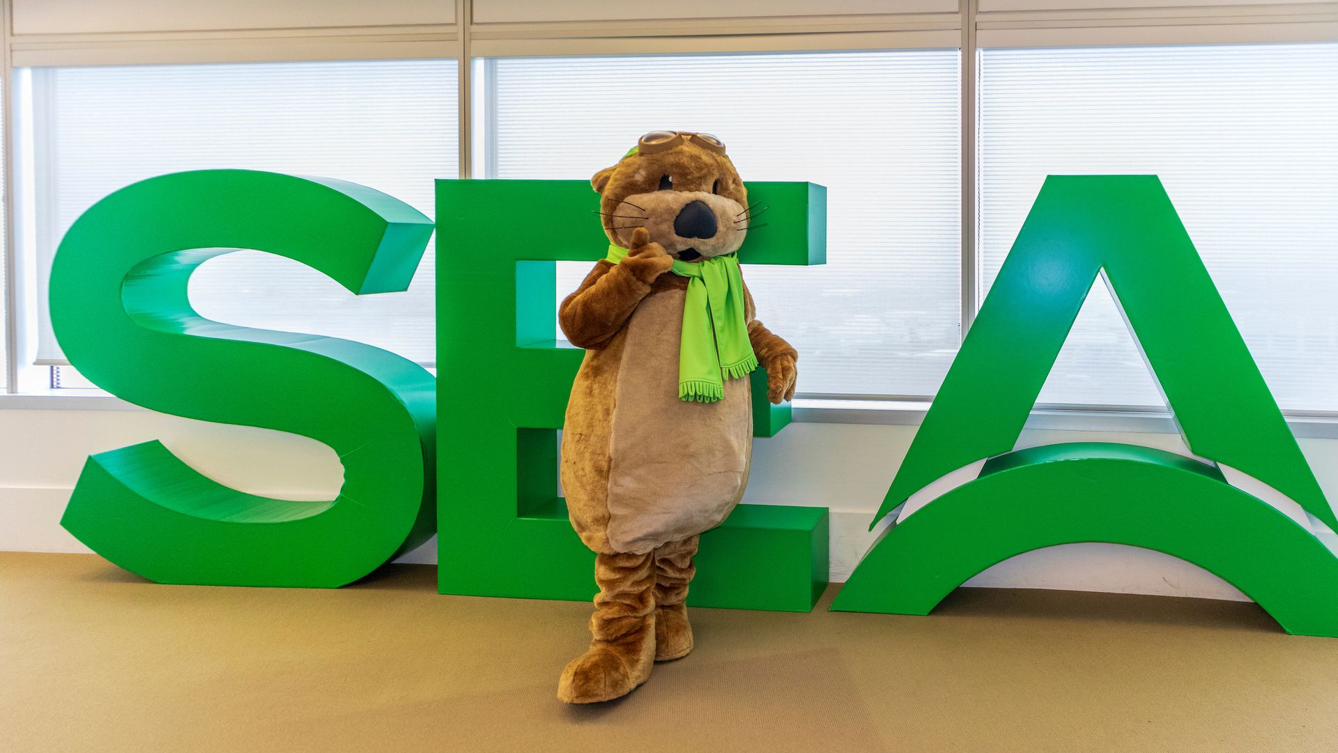 A person in a brown otter mascot costume with a green scarf and goggles stands indoors between large green letters S E and A.