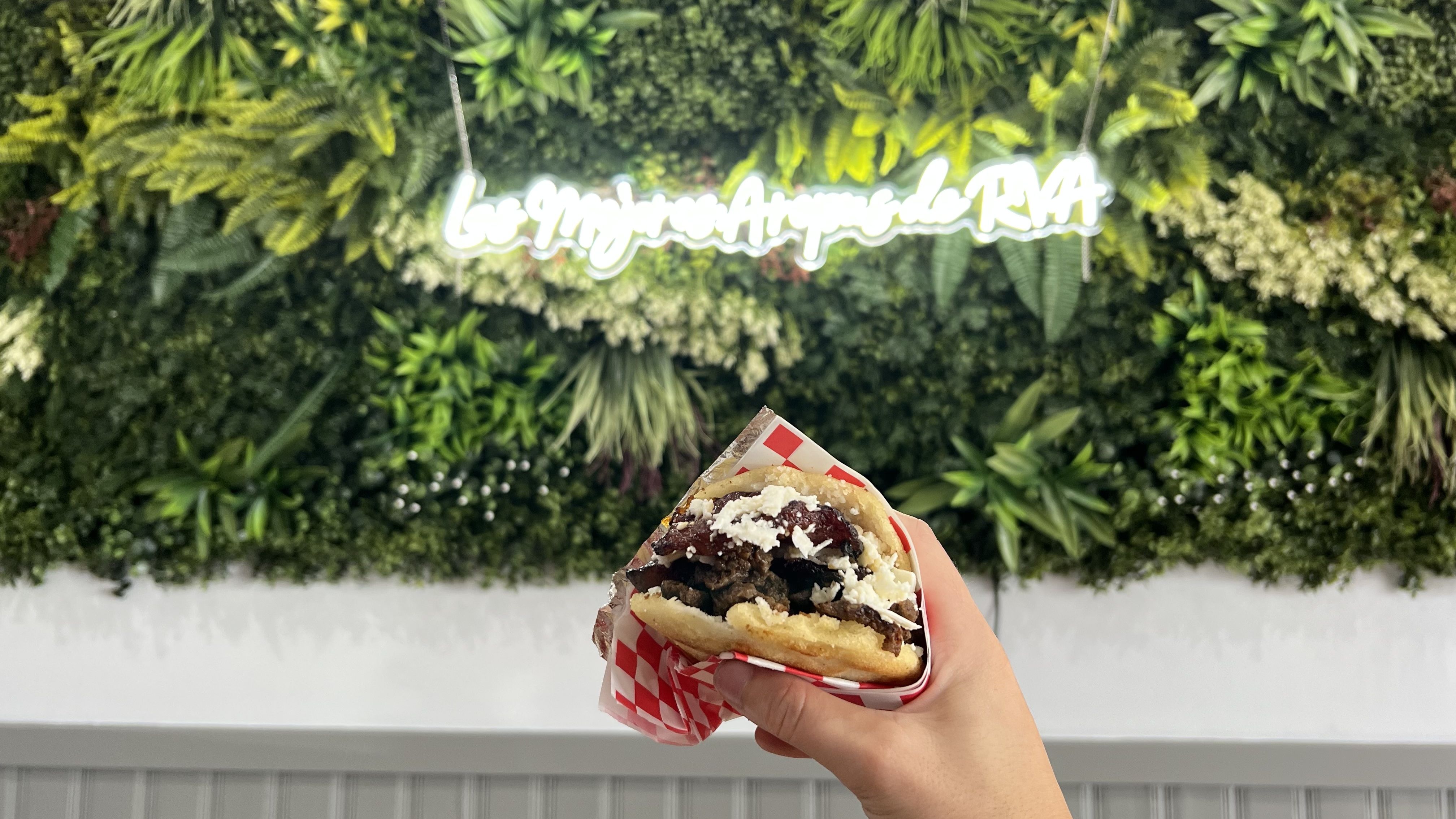 Hand holding arepa filled with meat and white cheese, wrapped in red and white paper, with a green leafy wall and blurred neon sign saying "Los mejores arepas de RVA" in background.
