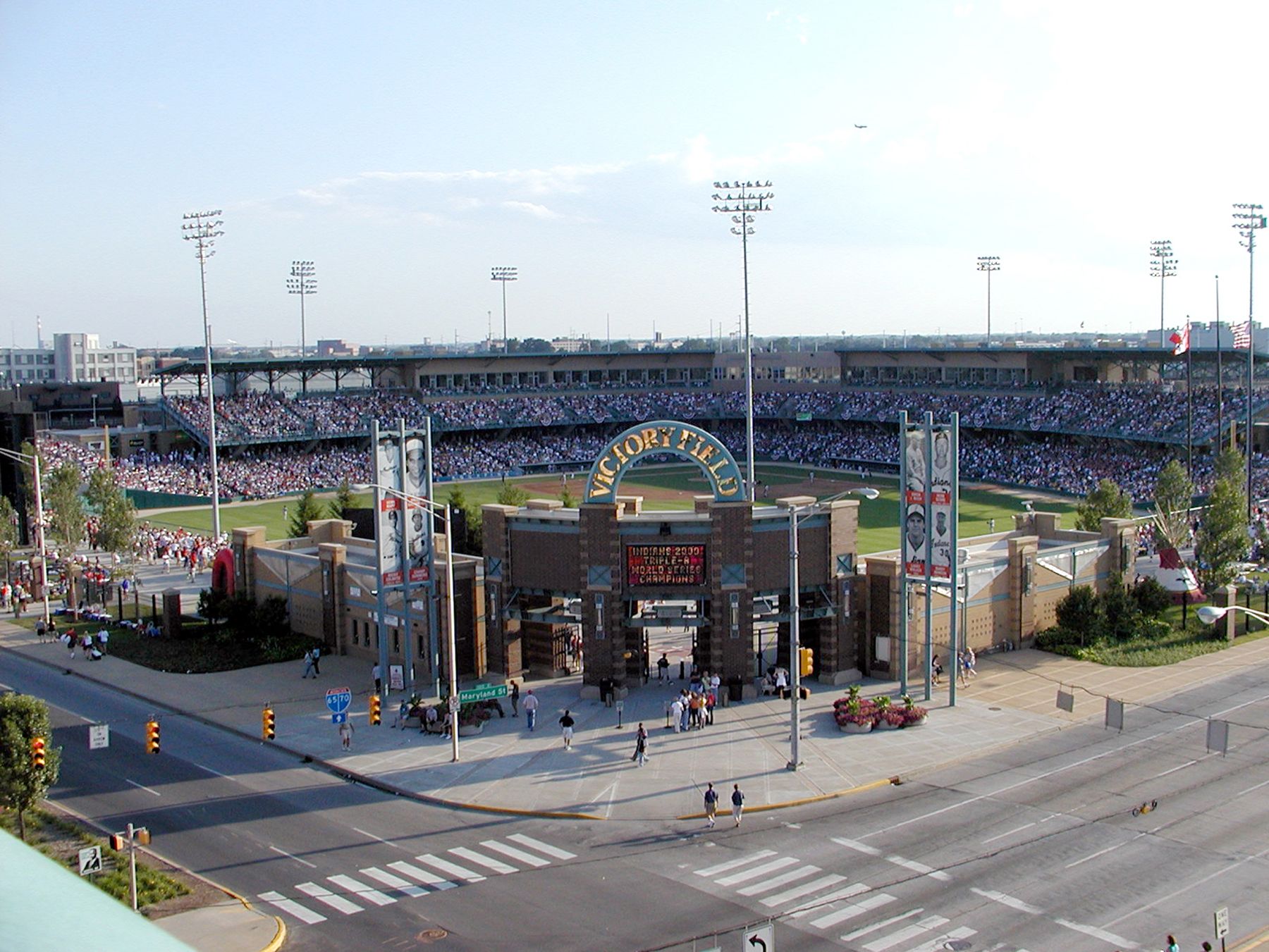 Wide view of "Victory Field" stadium filled with fans; tall light towers, brick gate arch reading "VICTORY FIELD"; busy street and crosswalk in the foreground under a clear sky.
