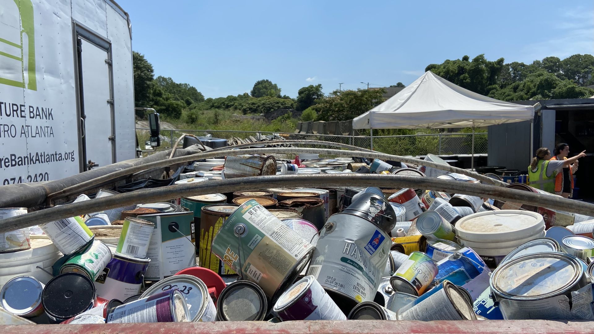 Truck bed filled with paint cans and scrap metal; a white trailer with partial text on the left. In the background, a white canopy tent, two workers in safety vests, blue sky and trees.
